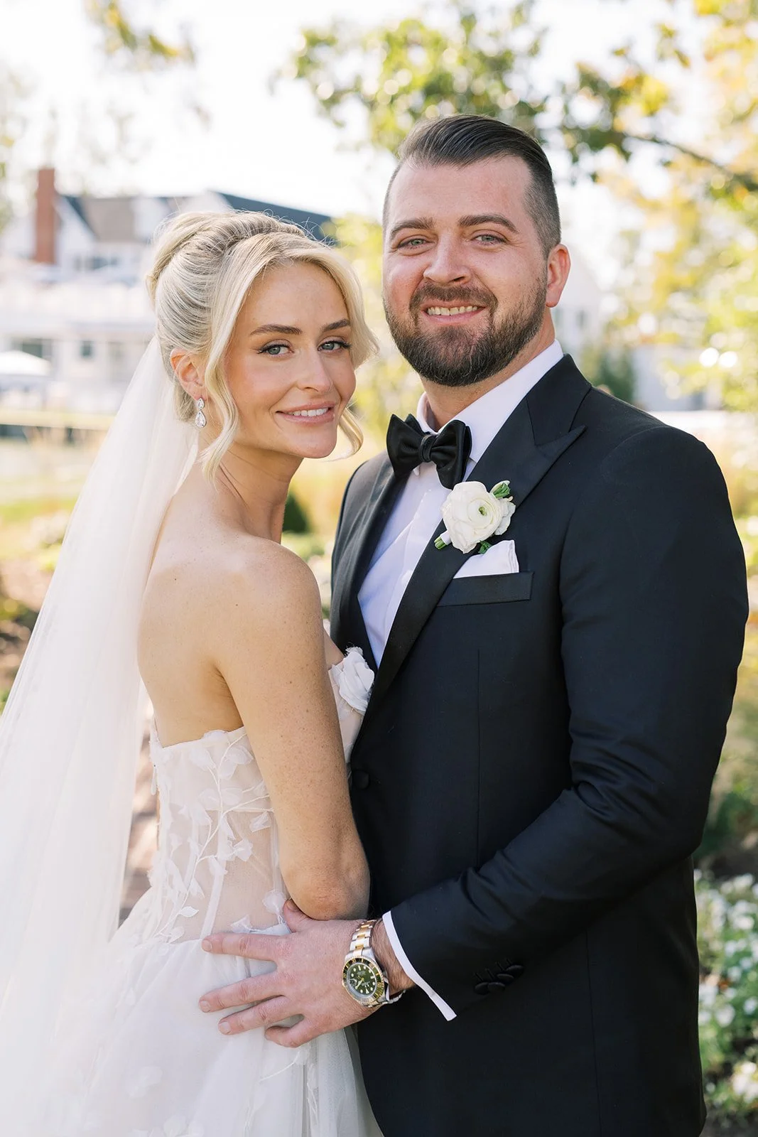 A bride and groom posing together outdoors on their wedding day, smiling at the camera. The bride is wearing a strapless wedding gown with a veil, and the groom is in a black tuxedo with a black bow tie and a white boutonniere.