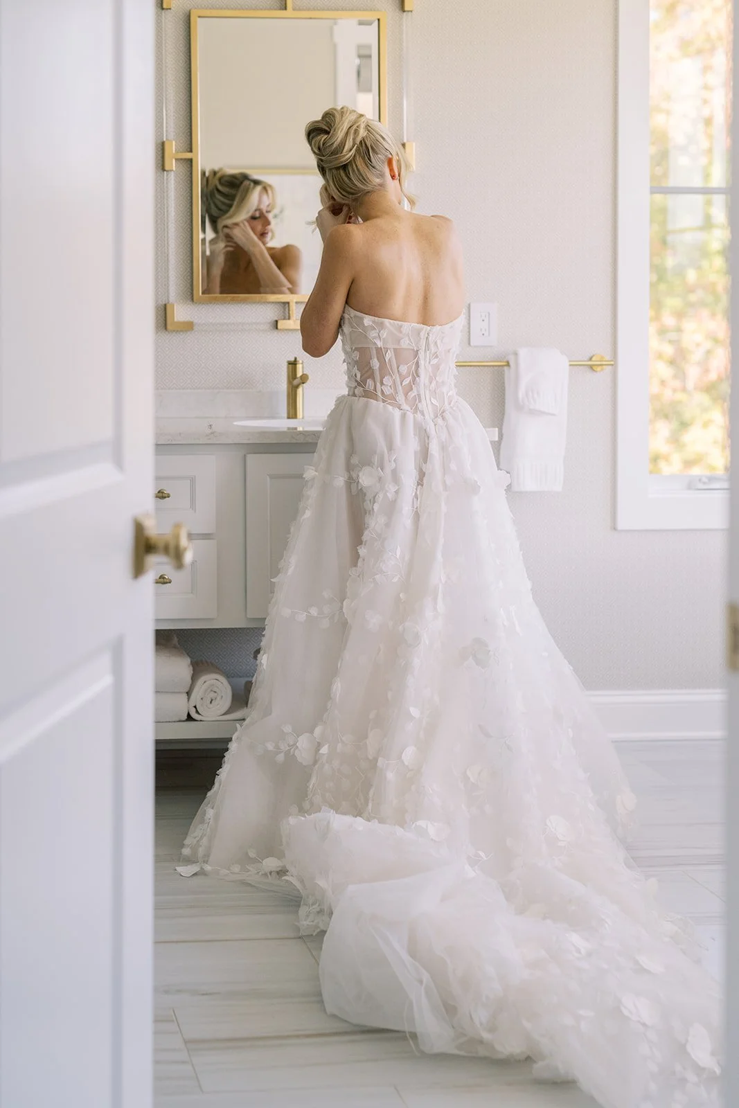 A woman in a strapless white wedding dress with floral lace details stands in front of a mirror in a bright bathroom, adjusting her hair.