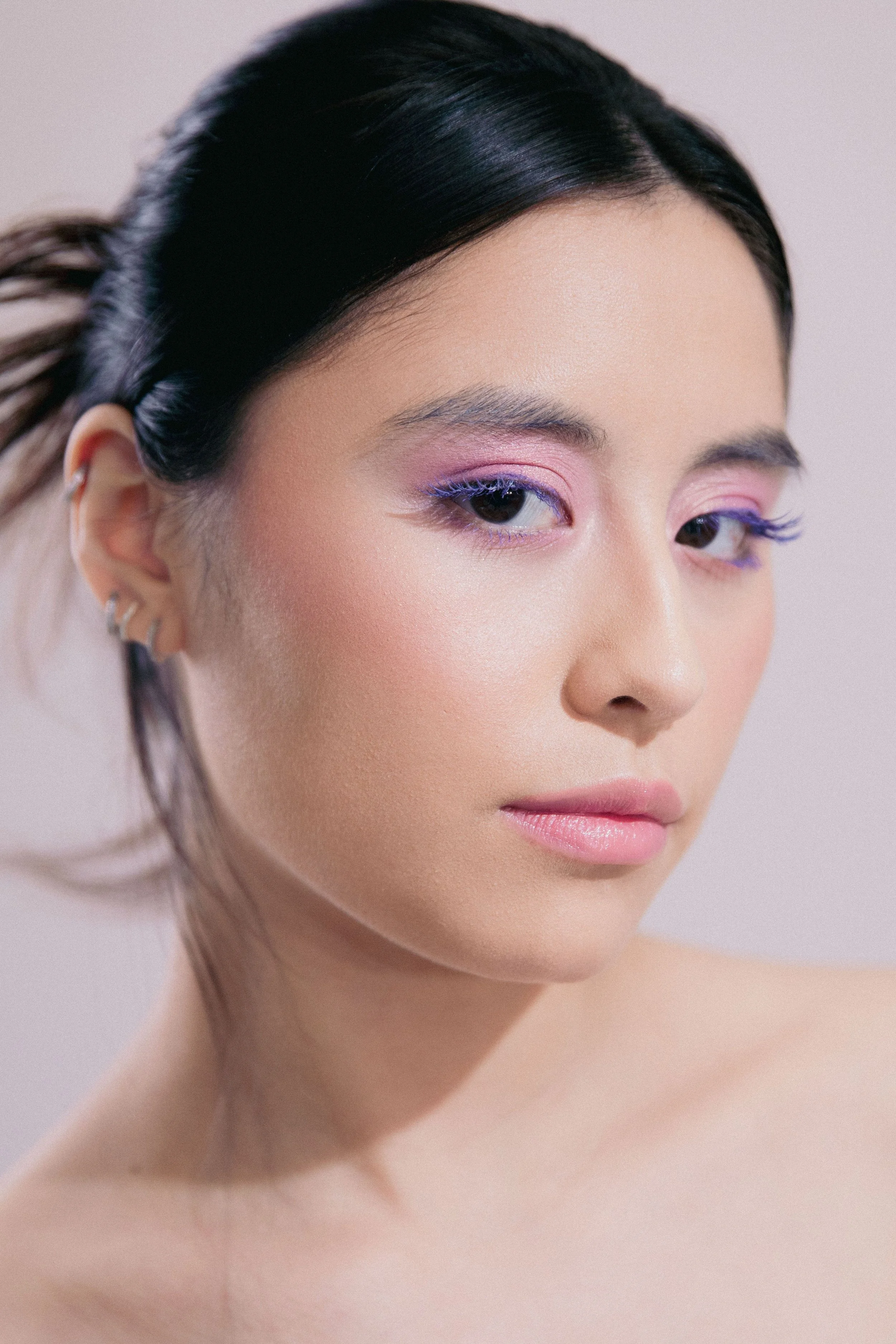 A close-up portrait of a woman with dark hair, light makeup, and colorful eyeshadow in pink and purple hues, looking slightly to her right with a neutral expression.