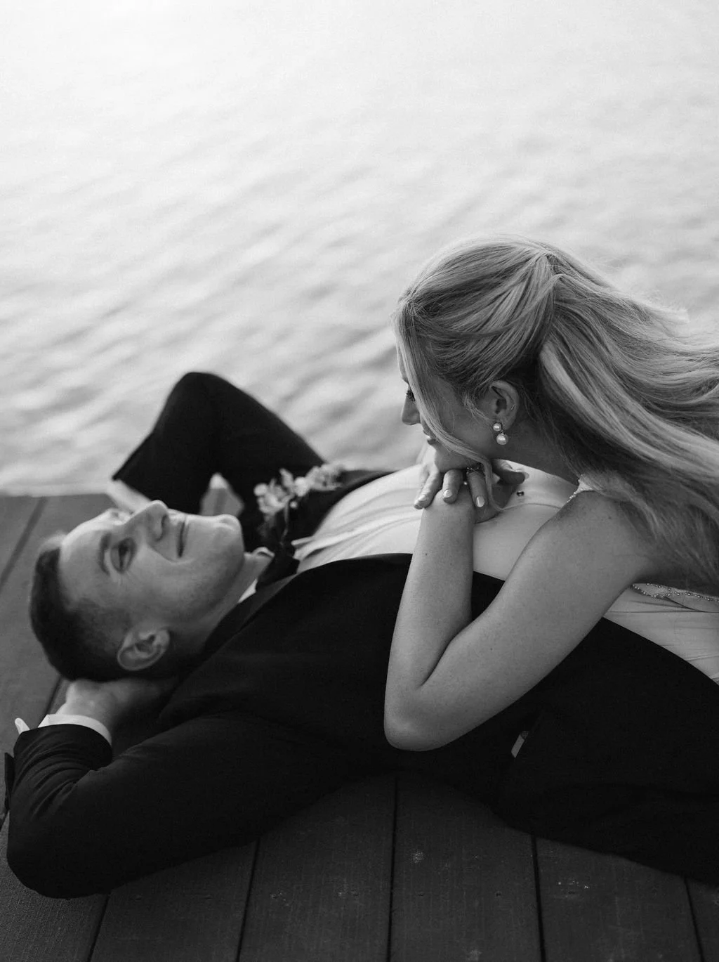 A black-and-white photo of a couple lying on a dock by a body of water. The man is lying on his back with his head resting on his hand, and the woman is leaning over him, looking at him affectionately.
