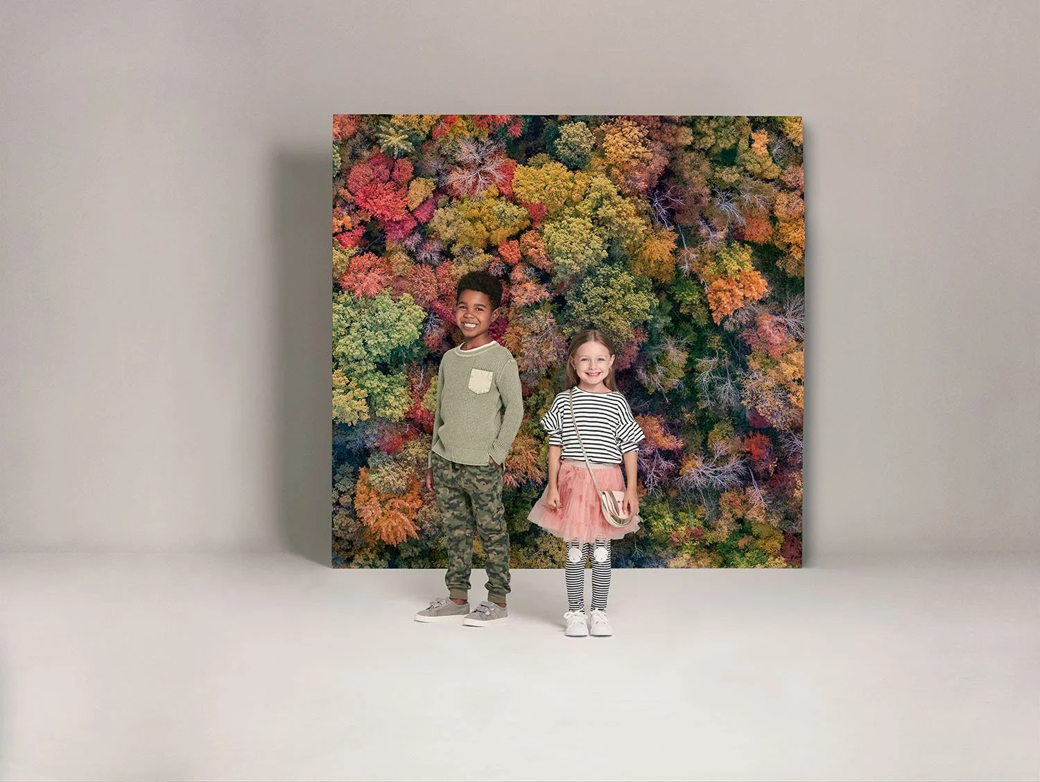 Two children standing in front of a large colorful autumn leaf mural, smiling. The boy is wearing a light green sweater and camouflage pants, while the girl is dressed in a striped top, pink tulle skirt, and striped tights, holding a small bag.