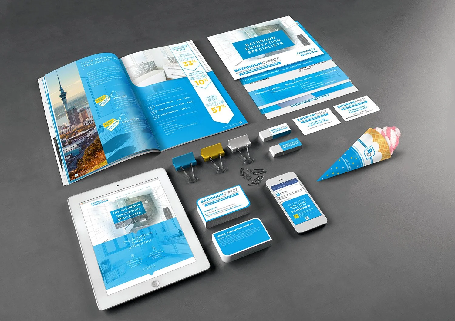 Marketing materials and branding items for Bathroom Direct, including brochures, flyers, business cards, branded ice cream cone, tablet, smartphone, paper clips, binder clips, and colored clothespins, all arranged on a gray surface.