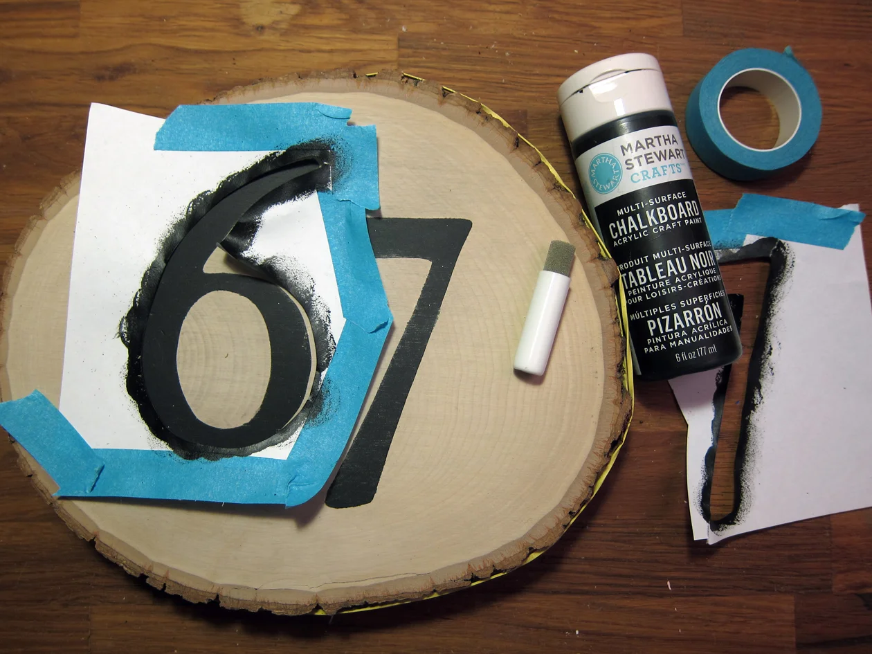 Create a number stencil. Pick a font and print out numbers to a piece of paper in the appropriate size. Cut numbers out with utility knife on a cutting mat. Save all number elements (i.e. inside circle of number 6). Position and center stencil to front of basswood round. Secure with tape. With paint and a foam pouncer, stencil the number. Remove paper stencil and let dry.