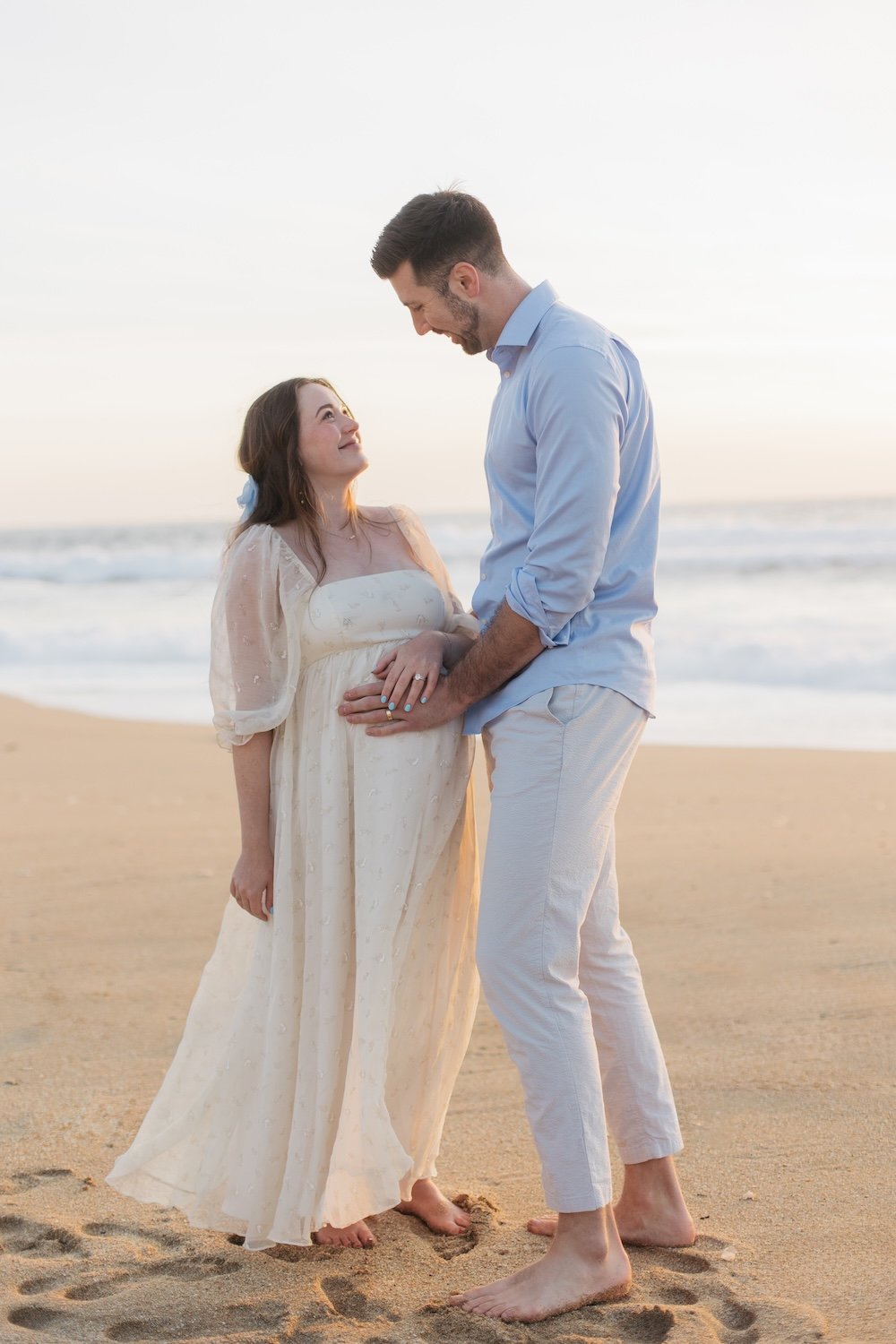 A pregnant woman and a man are standing barefoot on the beach at sunset, looking at each other lovingly, with the ocean in the background.