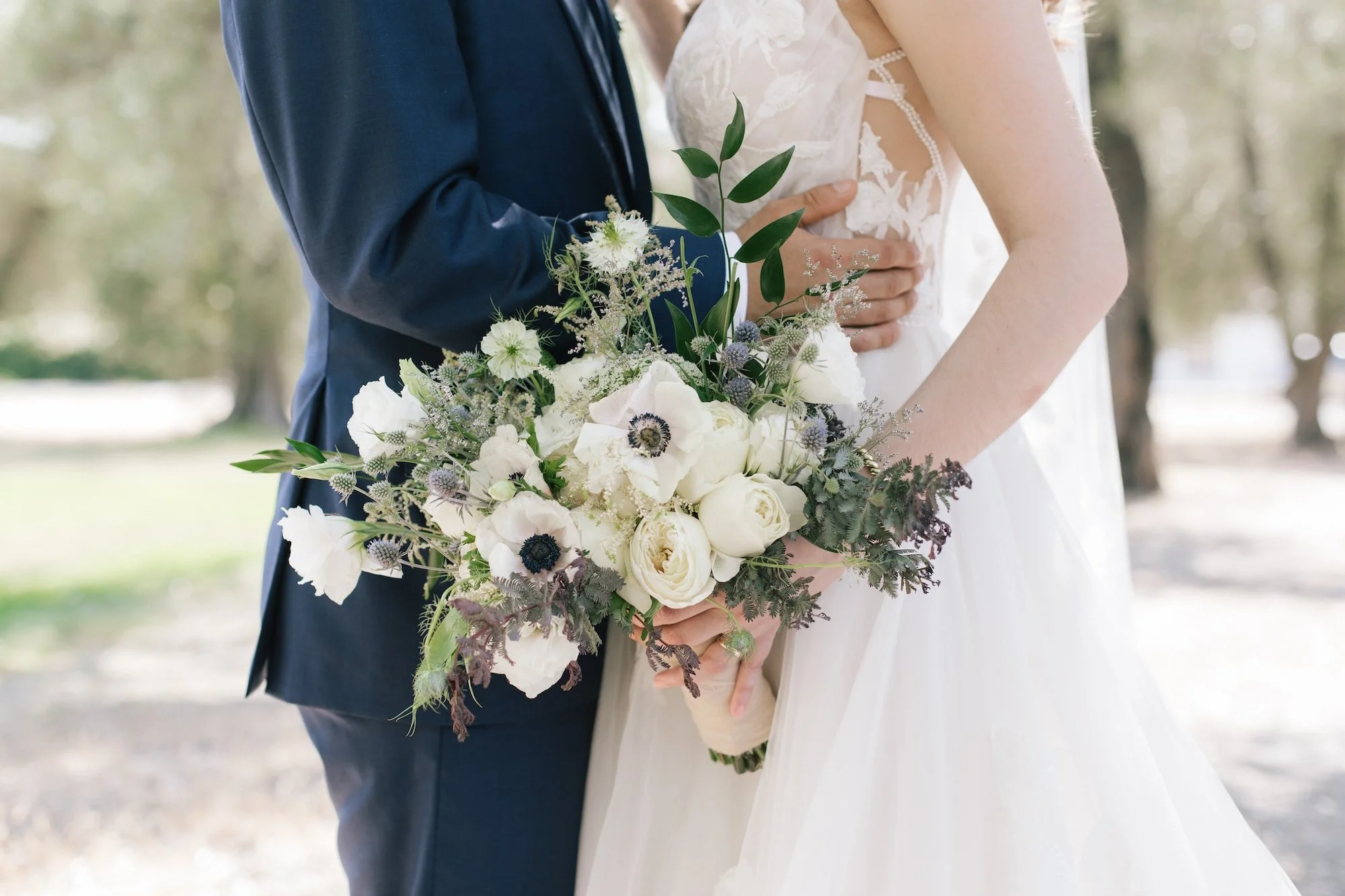 Close-up of a wedding couple with the groom in a navy suit and the bride in a white lace gown holding a bouquet of white roses, anemones, and greenery outdoors.