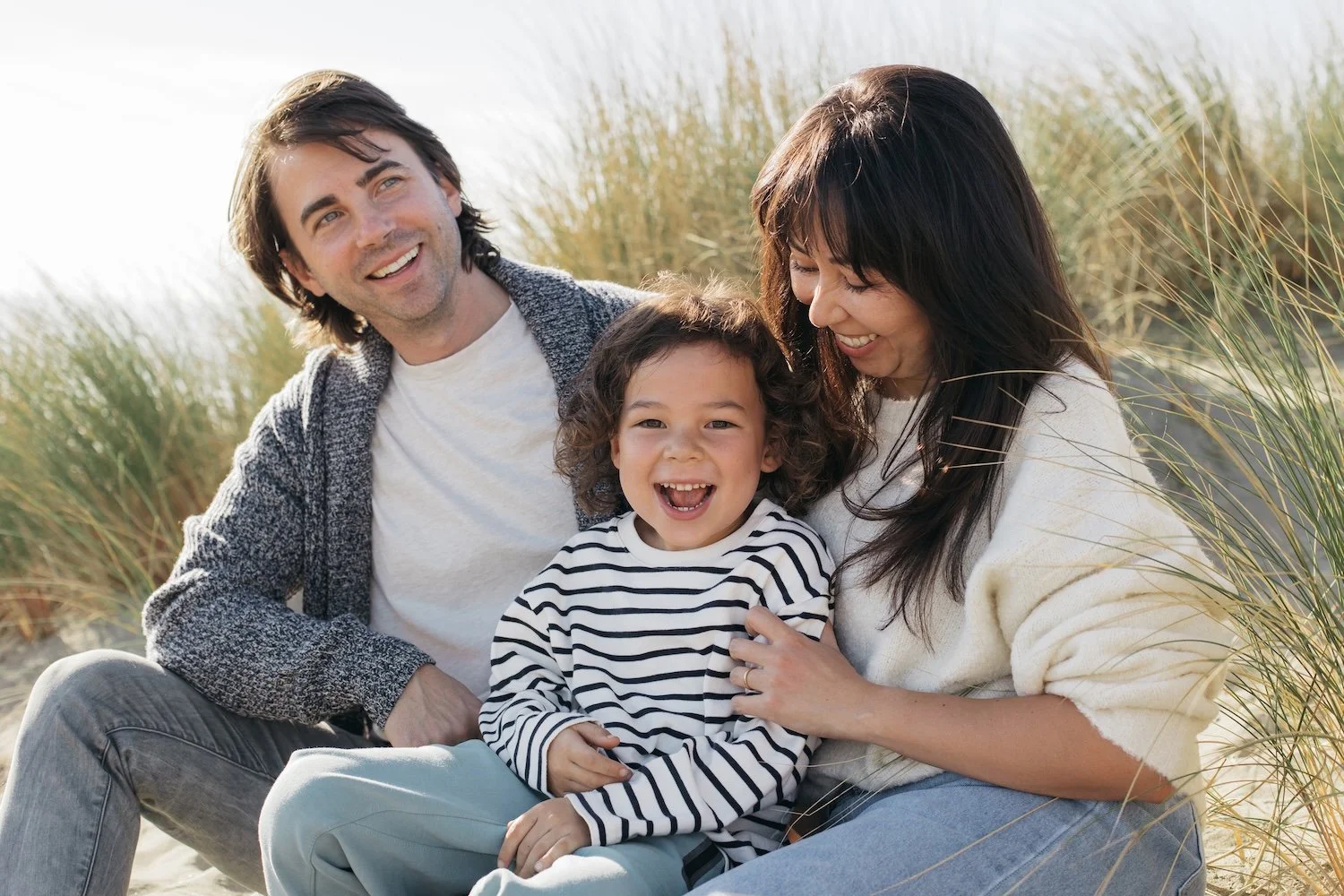 A happy family of three sitting outdoors near tall grass, smiling and laughing together on a sunny day.