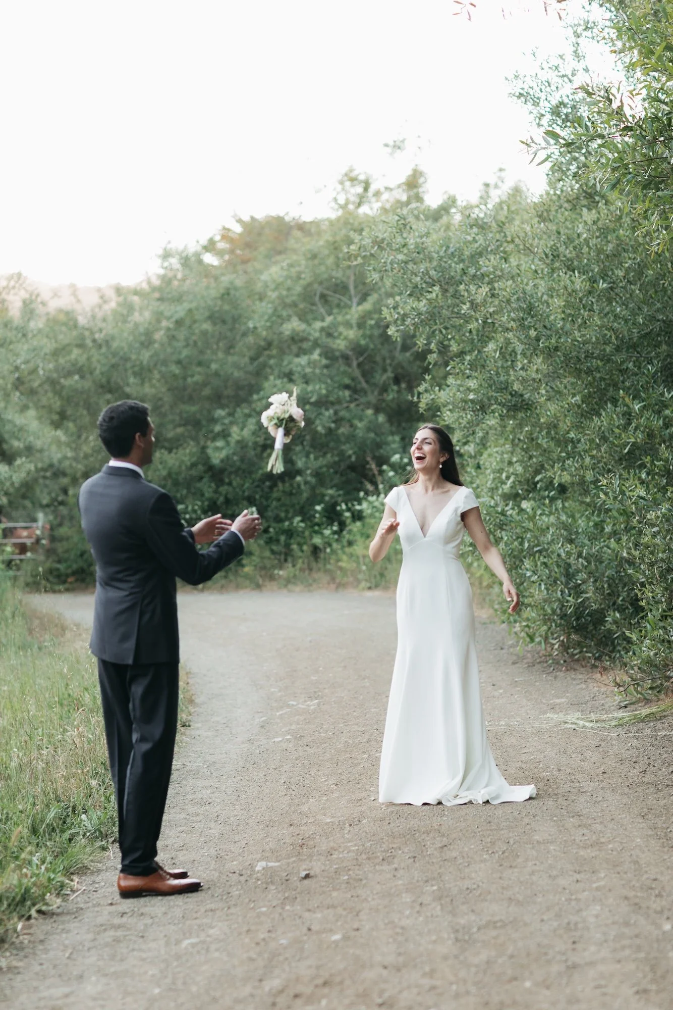 A bride in a white dress is laughing as a bouquet is thrown to her by a groom in a black suit, outdoors on a dirt path surrounded by green trees.