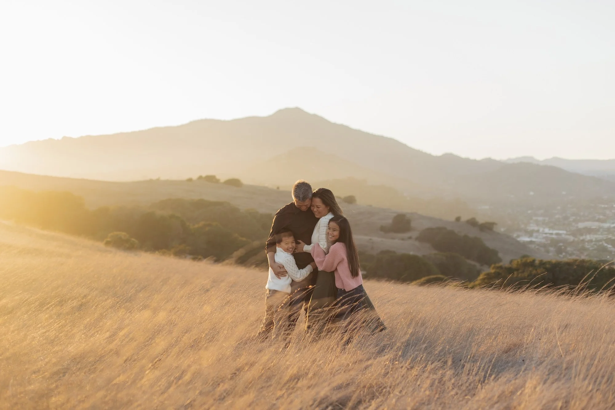 A family of four embracing each other in a golden field with mountains in the background at sunset.