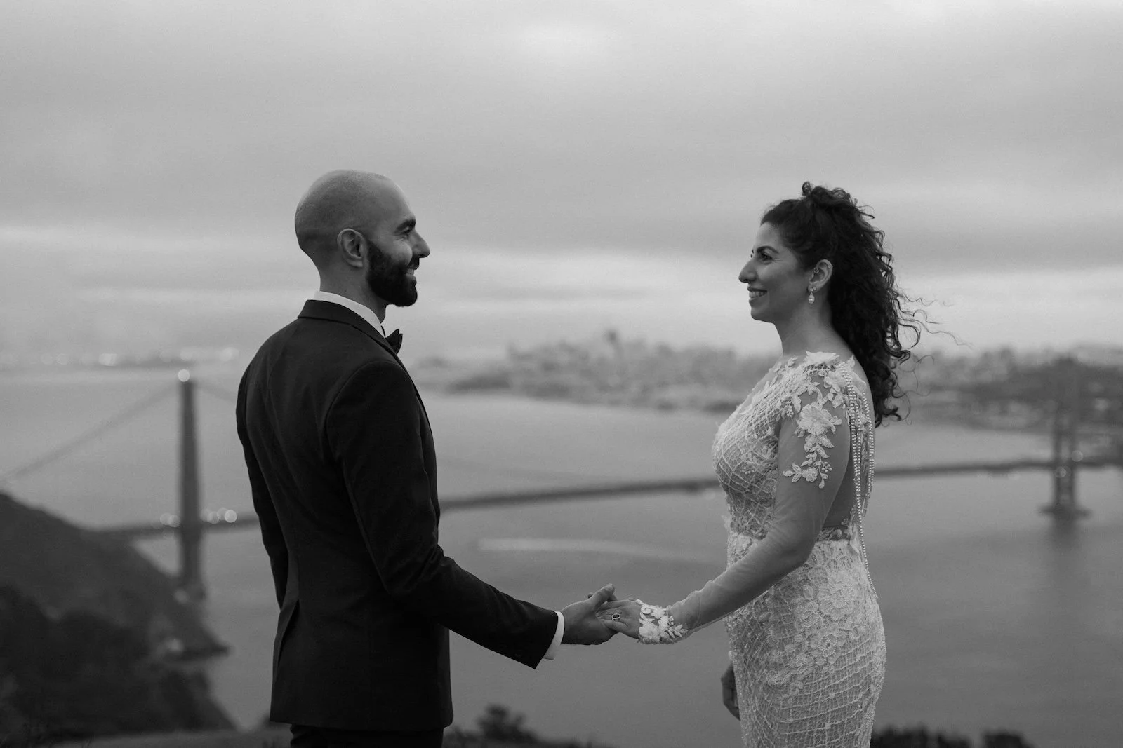 A black and white photo of a bride and groom holding hands and smiling at each other with the San Francisco Bay Bridge in the background.