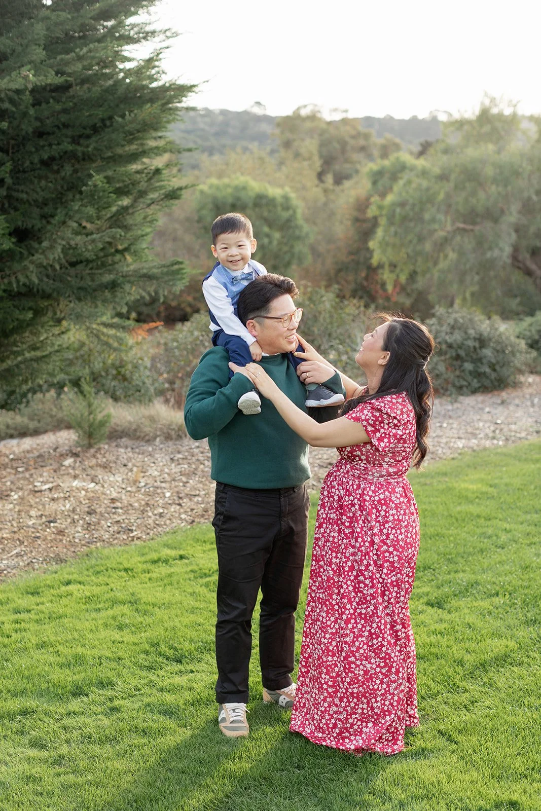 A family of three outdoors in a park, with a man carrying a smiling young boy on his shoulders and a woman in a pink floral dress touching the man's face, all smiling at each other.