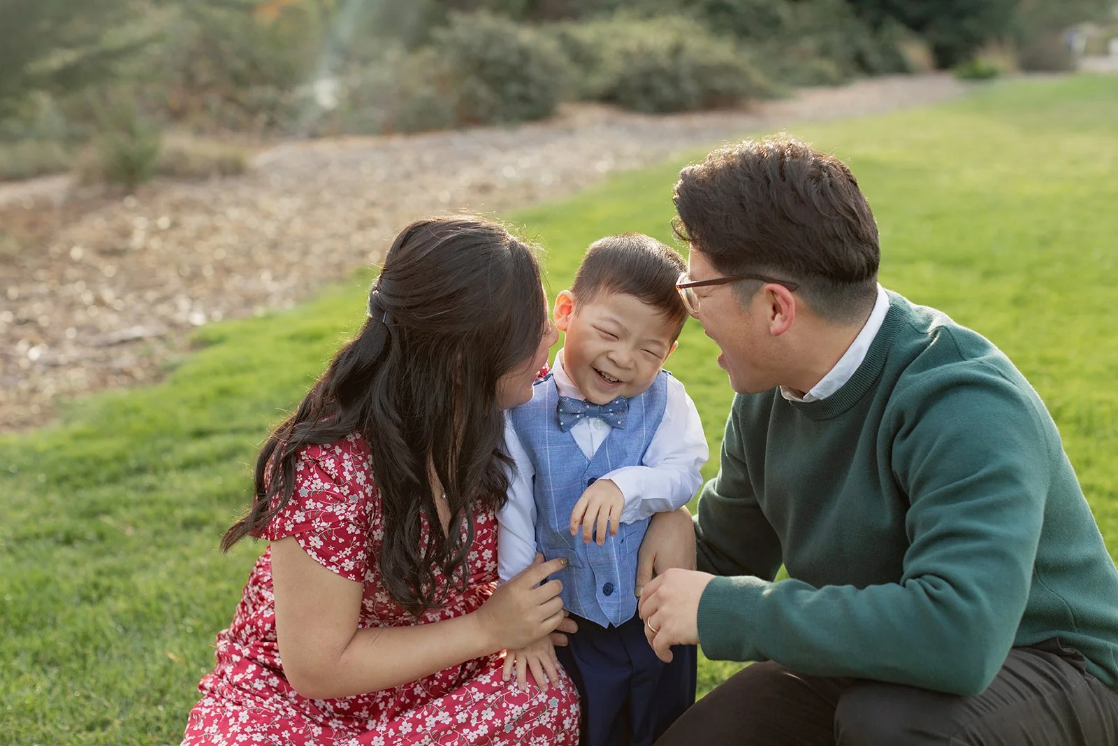 A happy Asian family consisting of a mother, father, and young son enjoying time together outdoors on a grassy field, smiling and laughing.