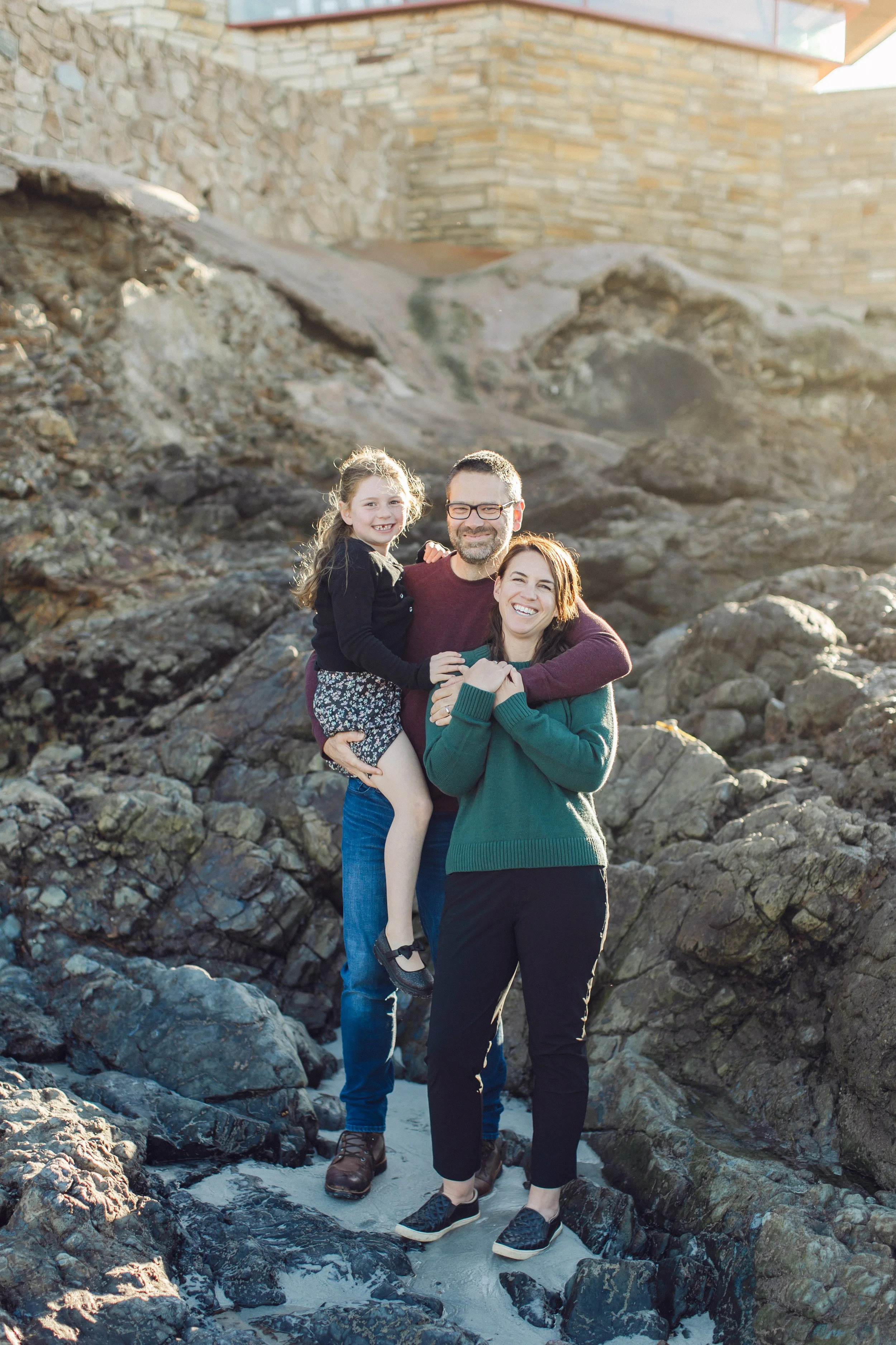 A family of three, a man, woman, and young girl, standing on rocks near the water's edge at a rocky beach, smiling and posing for the photo.