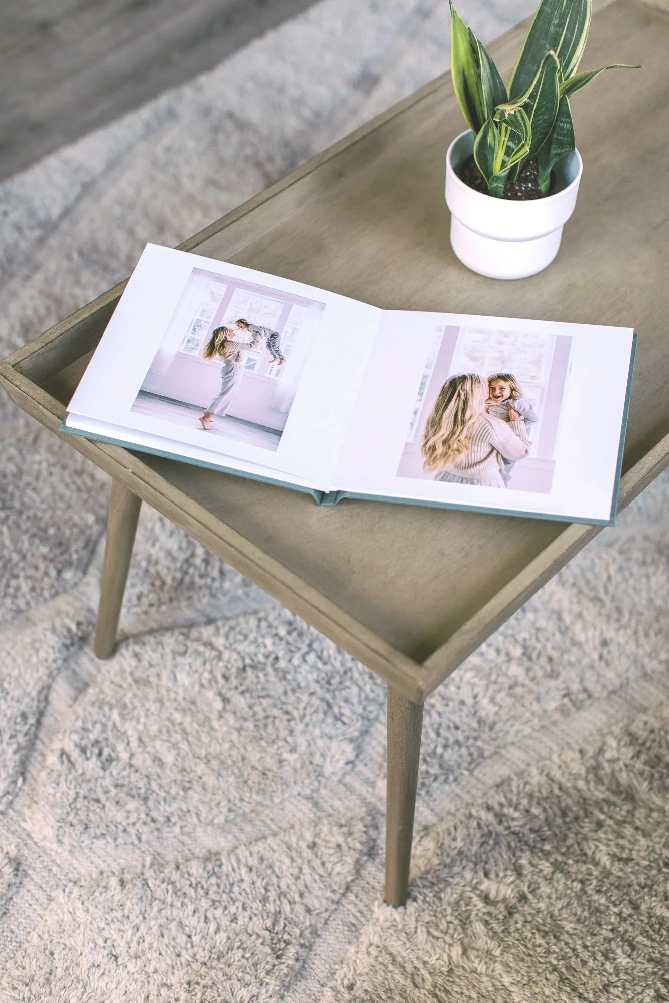 Open photo album with pictures of a woman and a young girl on a light wooden table, with a potted green plant nearby, on a light-colored carpeted floor.