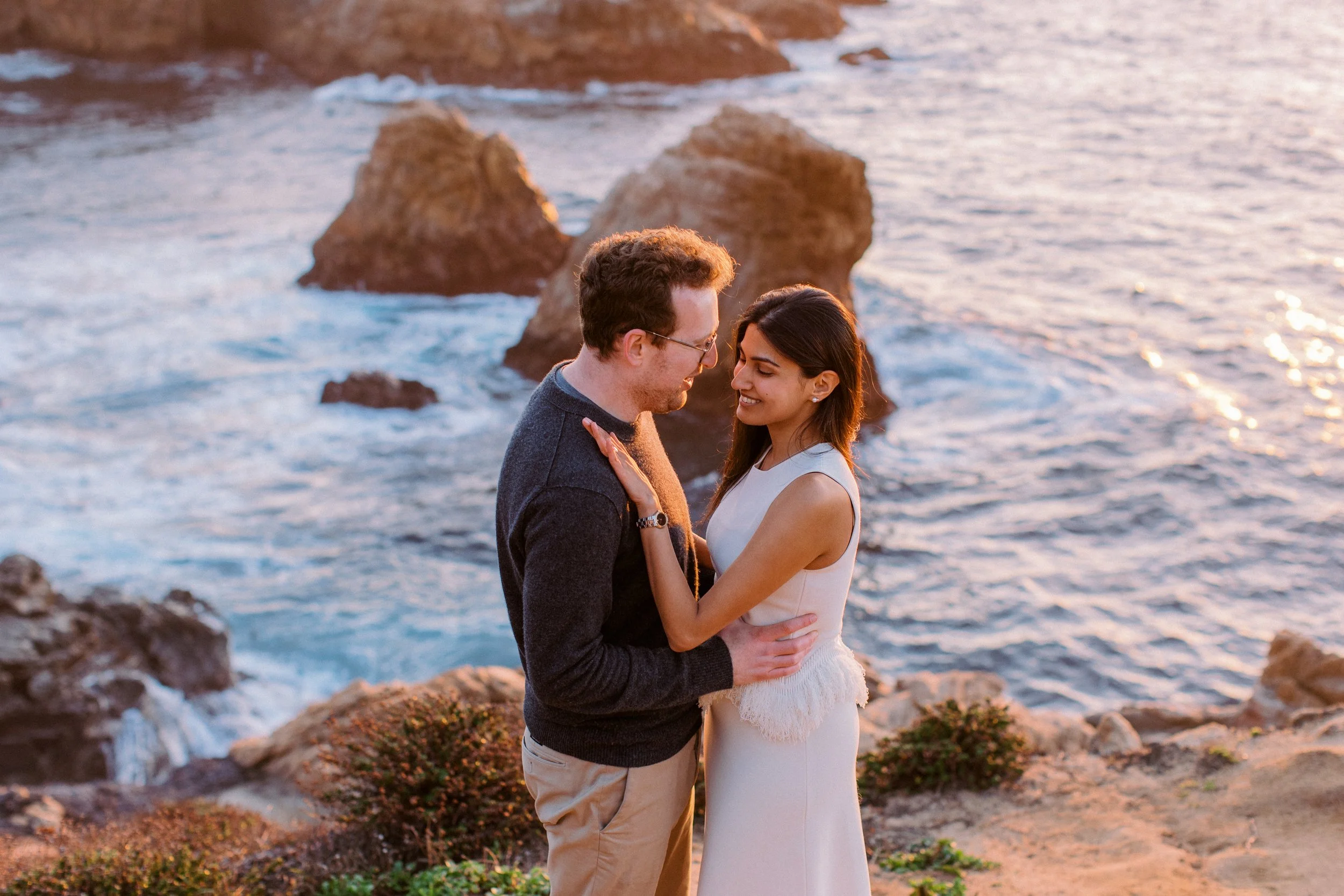 A couple stands close by the rocky shoreline at sunset, smiling at each other with the ocean waves behind them.