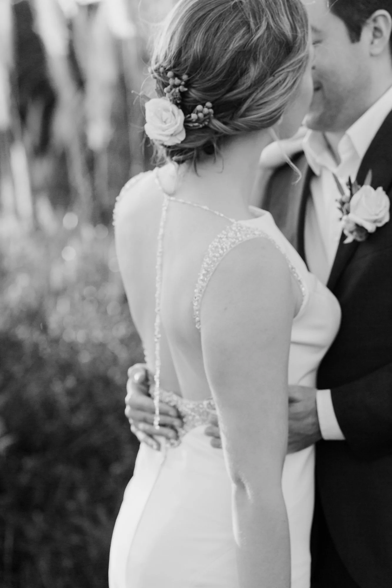 Black and white photo of a bride and groom sharing a close, intimate moment at their wedding, with the bride having floral hair accessories and wearing a sleeveless wedding dress, and the groom in a tuxedo with a boutonniere.