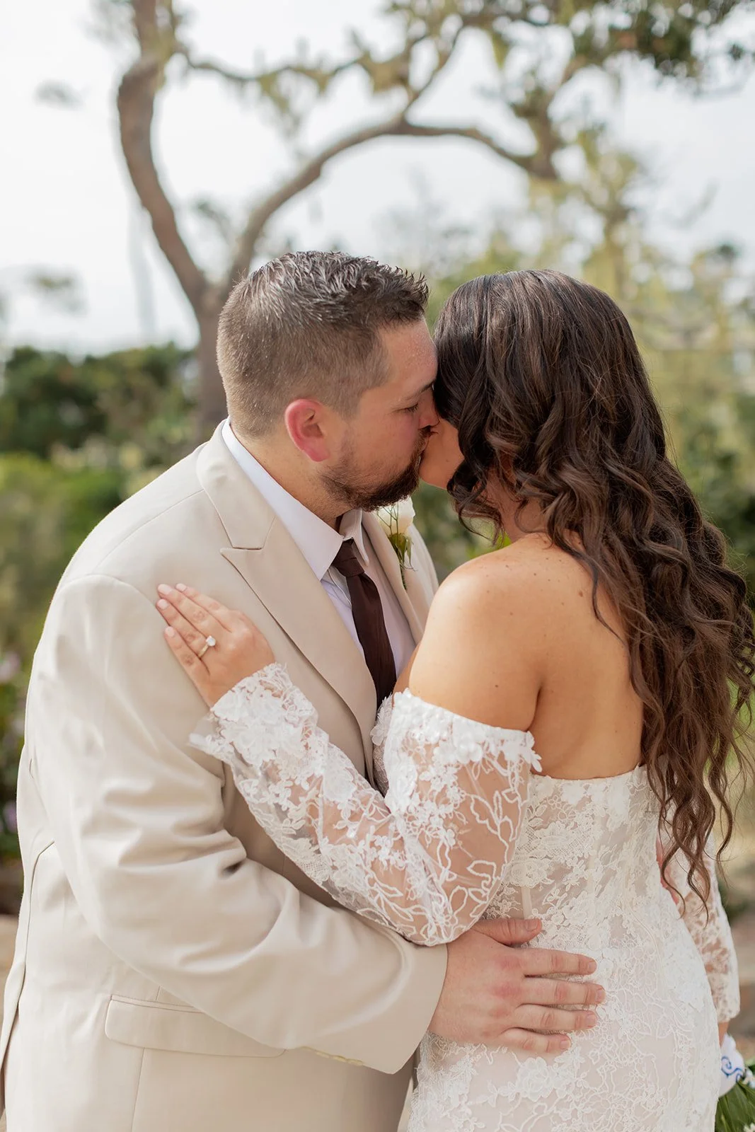 A bride and groom sharing a kiss during their wedding outdoors, with trees and a cloudy sky in the background.