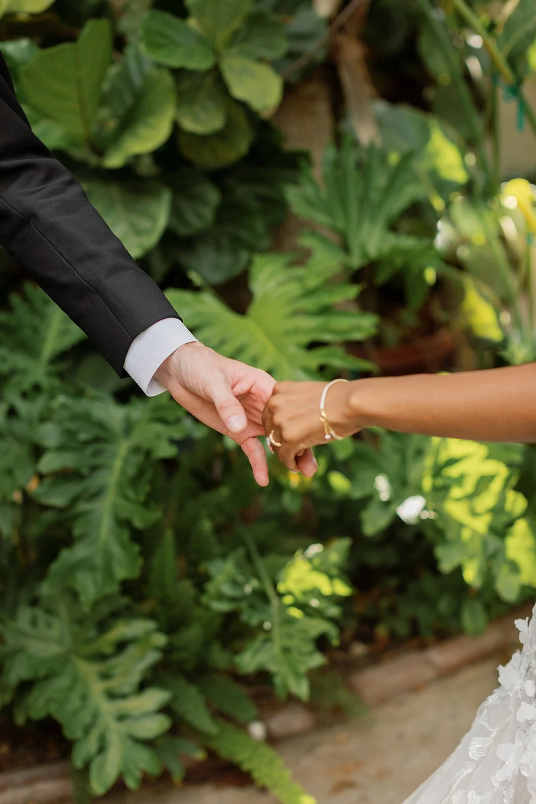 A couple holding hands, one in a black suit and the other in white dress, outdoors with lush green foliage in the background.