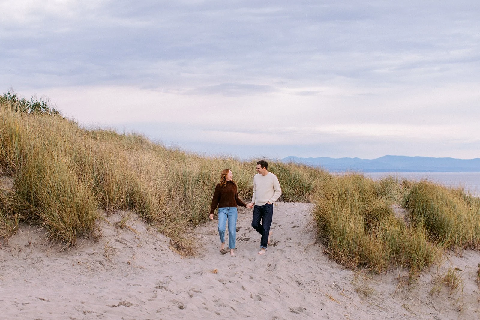 A couple walking hand in hand along a sandy beach path flanked by tall grass with the ocean and distant mountains in the background under a cloudy sky.