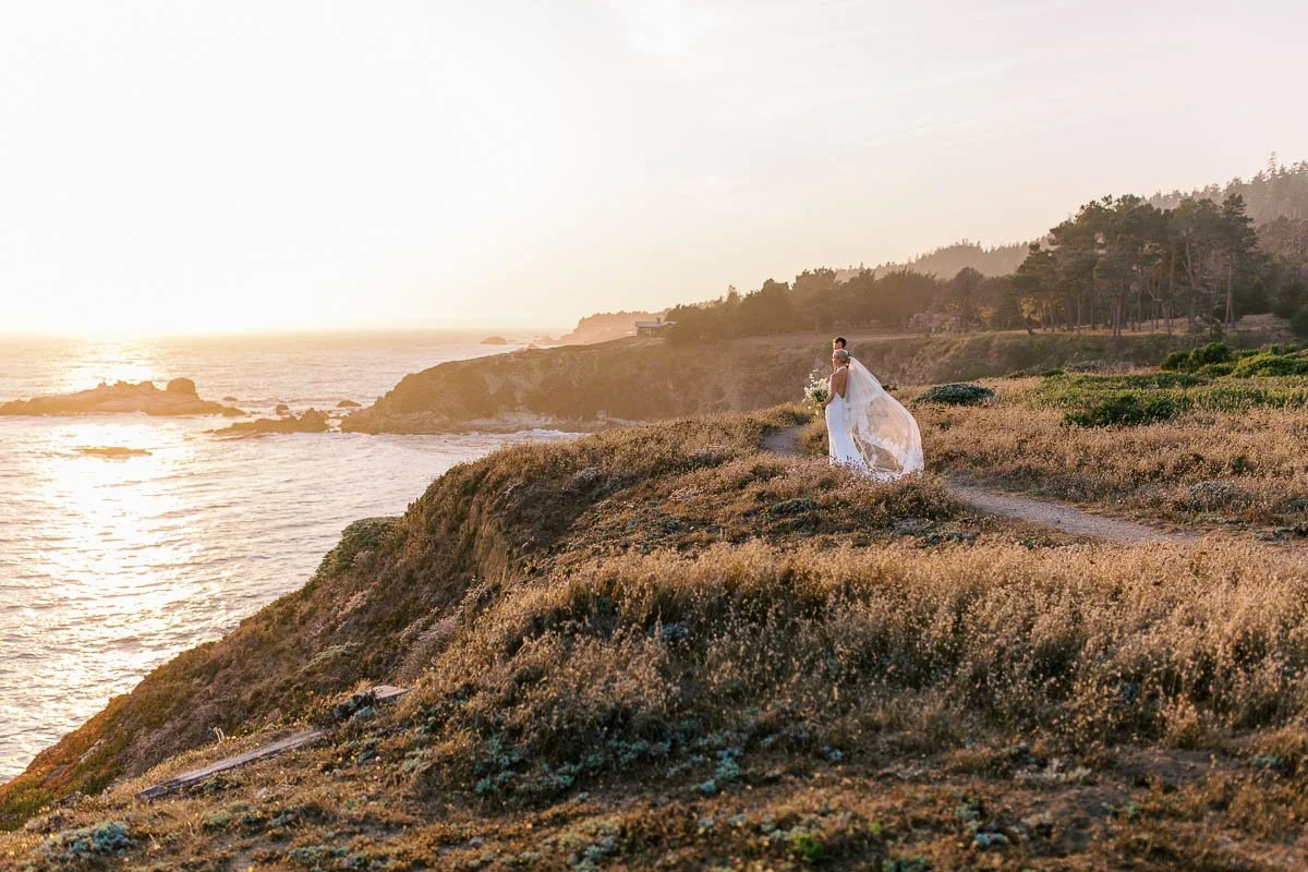 A bride in a white wedding dress holding a bouquet, standing on a cliffside trail overlooking the ocean during sunset.
