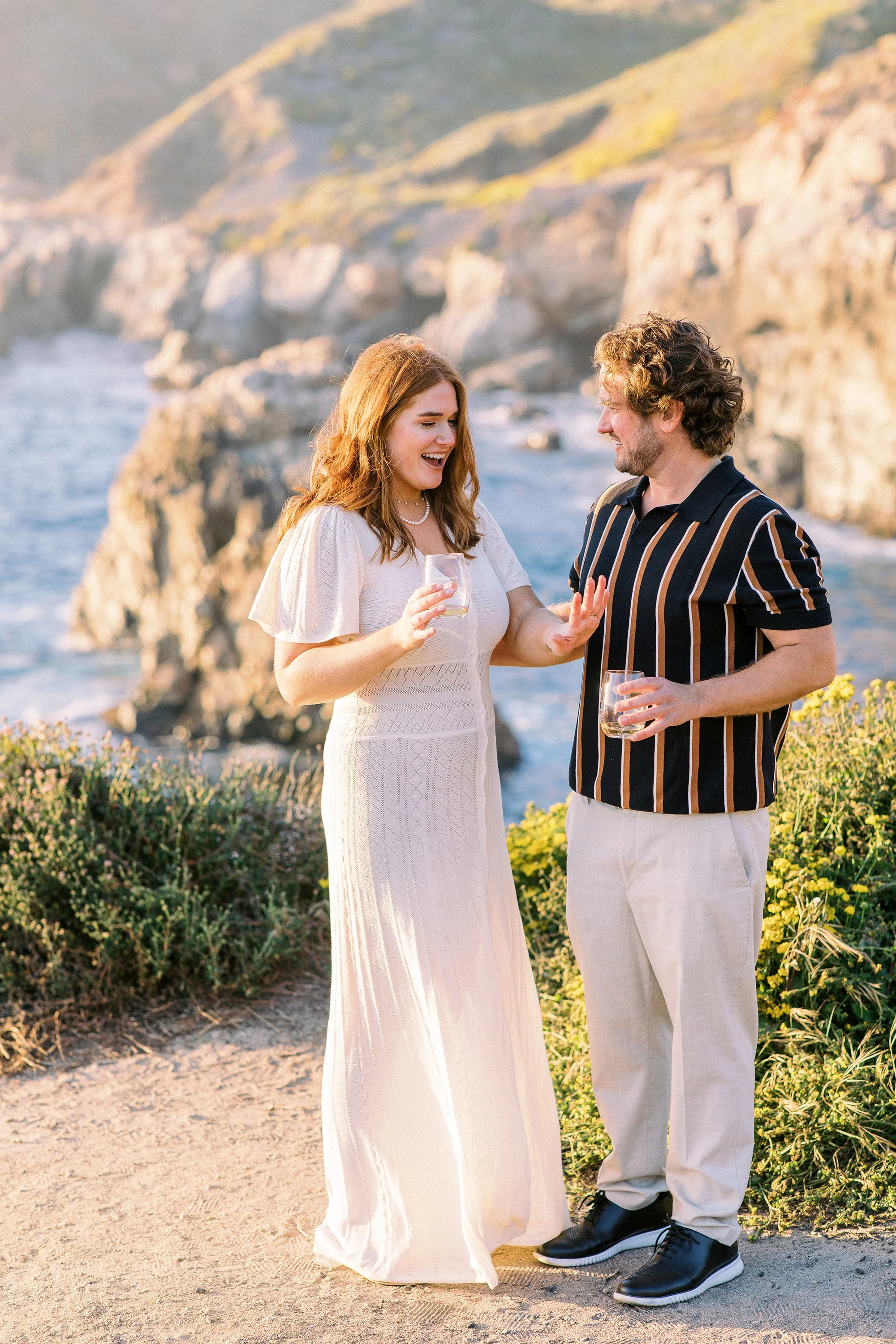 A woman and a man having a lively conversation, holding glasses of wine, outdoors near the coast with cliffs in the background.