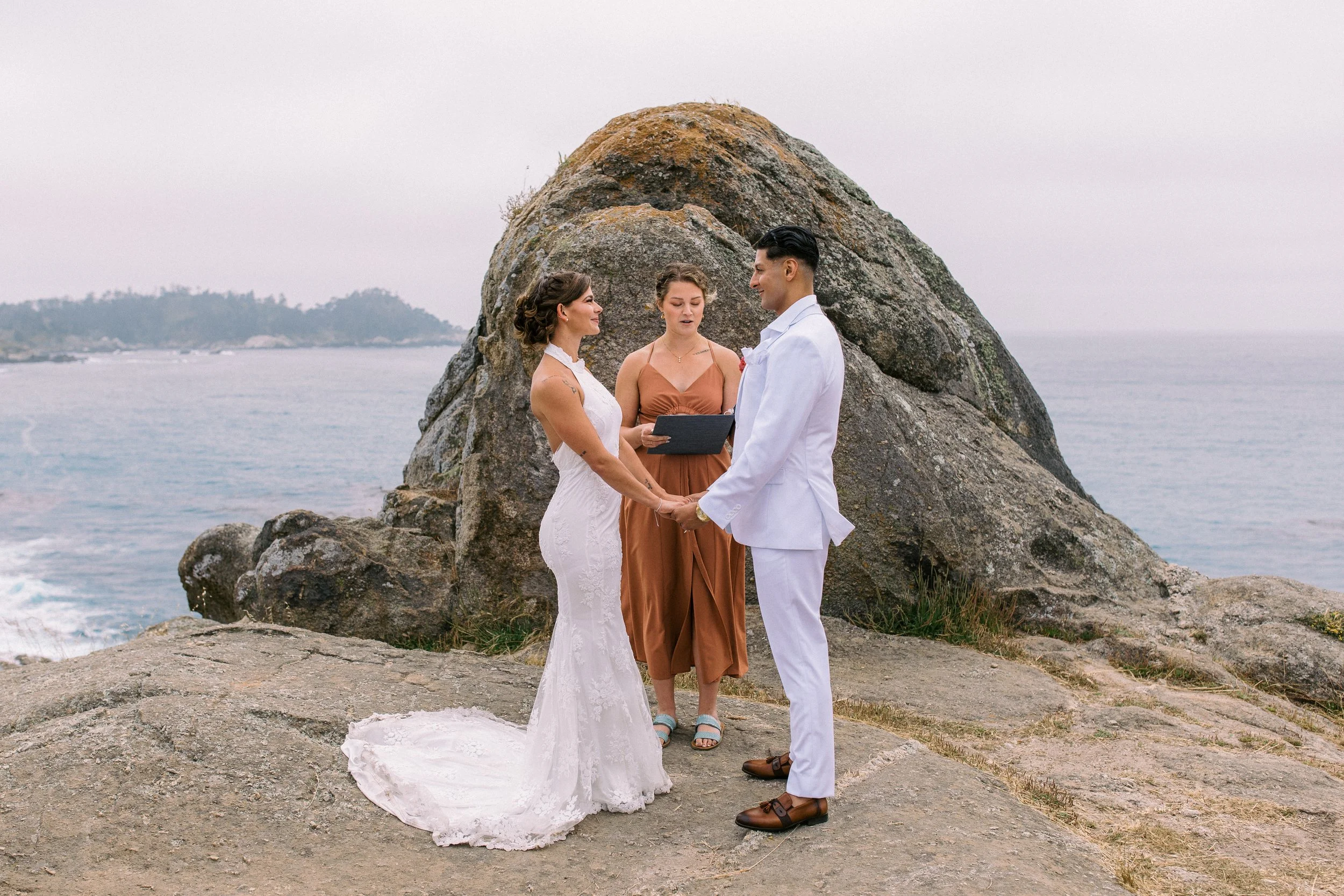 A wedding ceremony taking place outdoors on a rocky shore with an ocean and distant land in the background. A bride and groom are holding hands and facing each other, with a female officiant standing behind them, reading from a book.