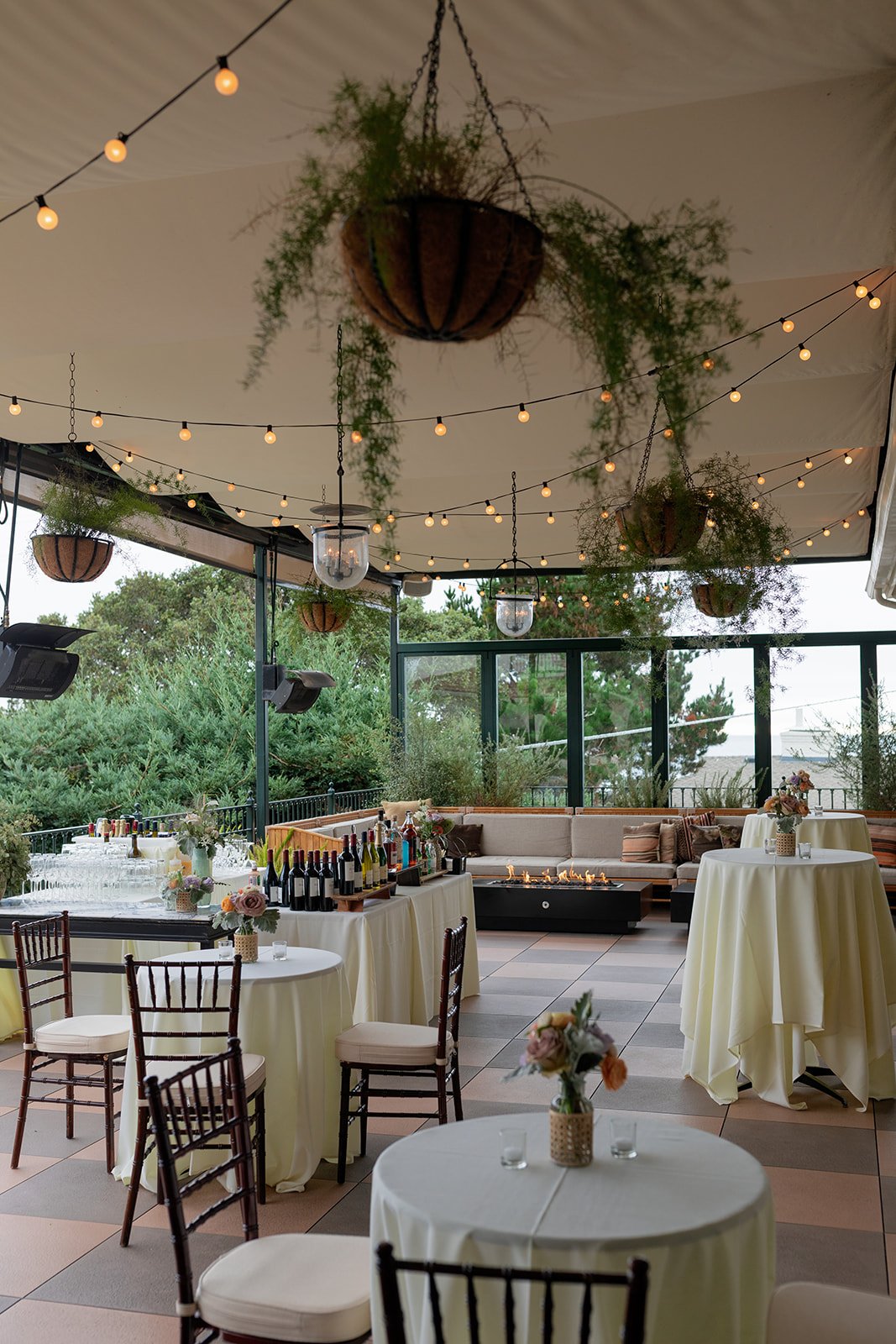 An outdoor event space decorated with hanging plants, string lights, and tables with white tablecloths and floral centerpieces, featuring a mix of chairs and a built-in seating area.