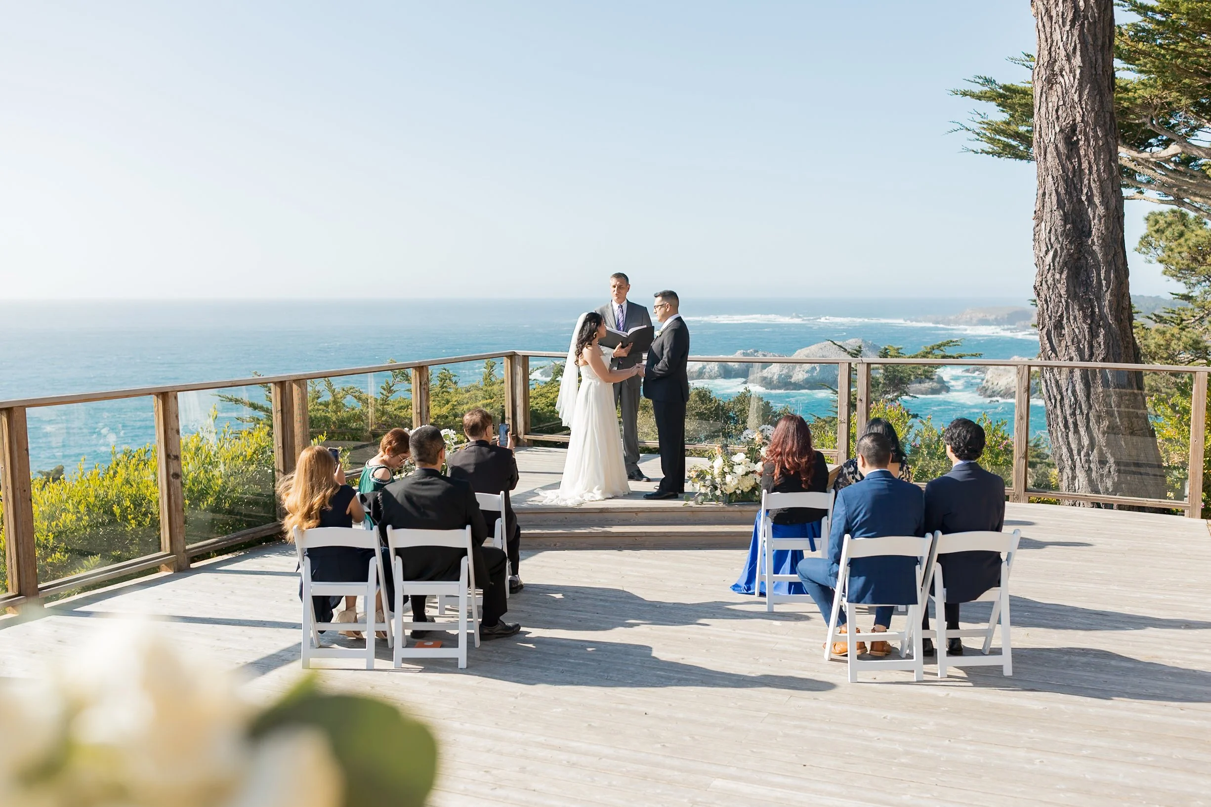 A wedding ceremony on a wooden terrace overlooking the ocean, with a bride and groom exchanging vows, officiant in background, and guests seated in white chairs.