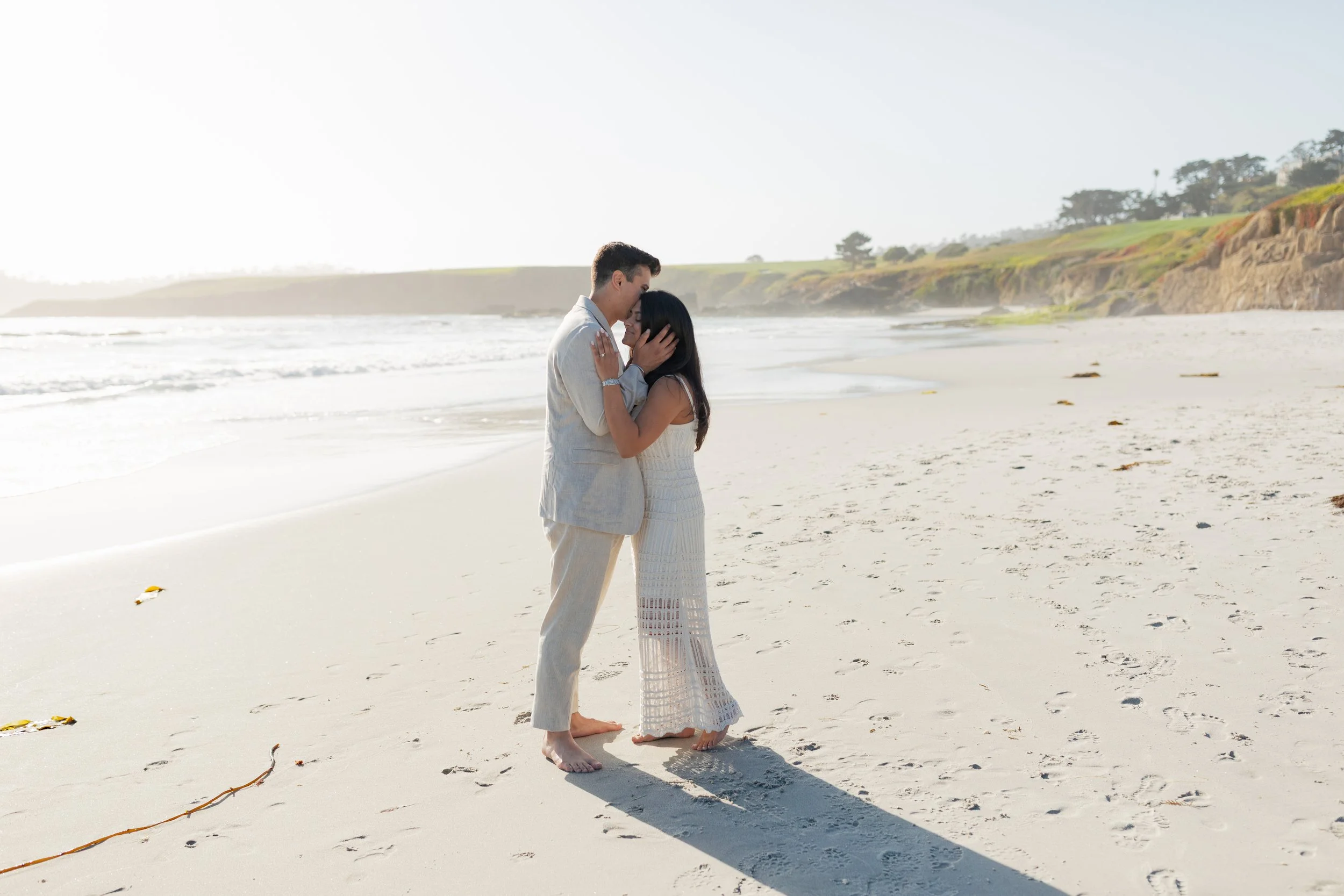 A couple standing closely on a sandy beach, sharing an intimate moment, with the ocean and cliffs in the background during the daytime.