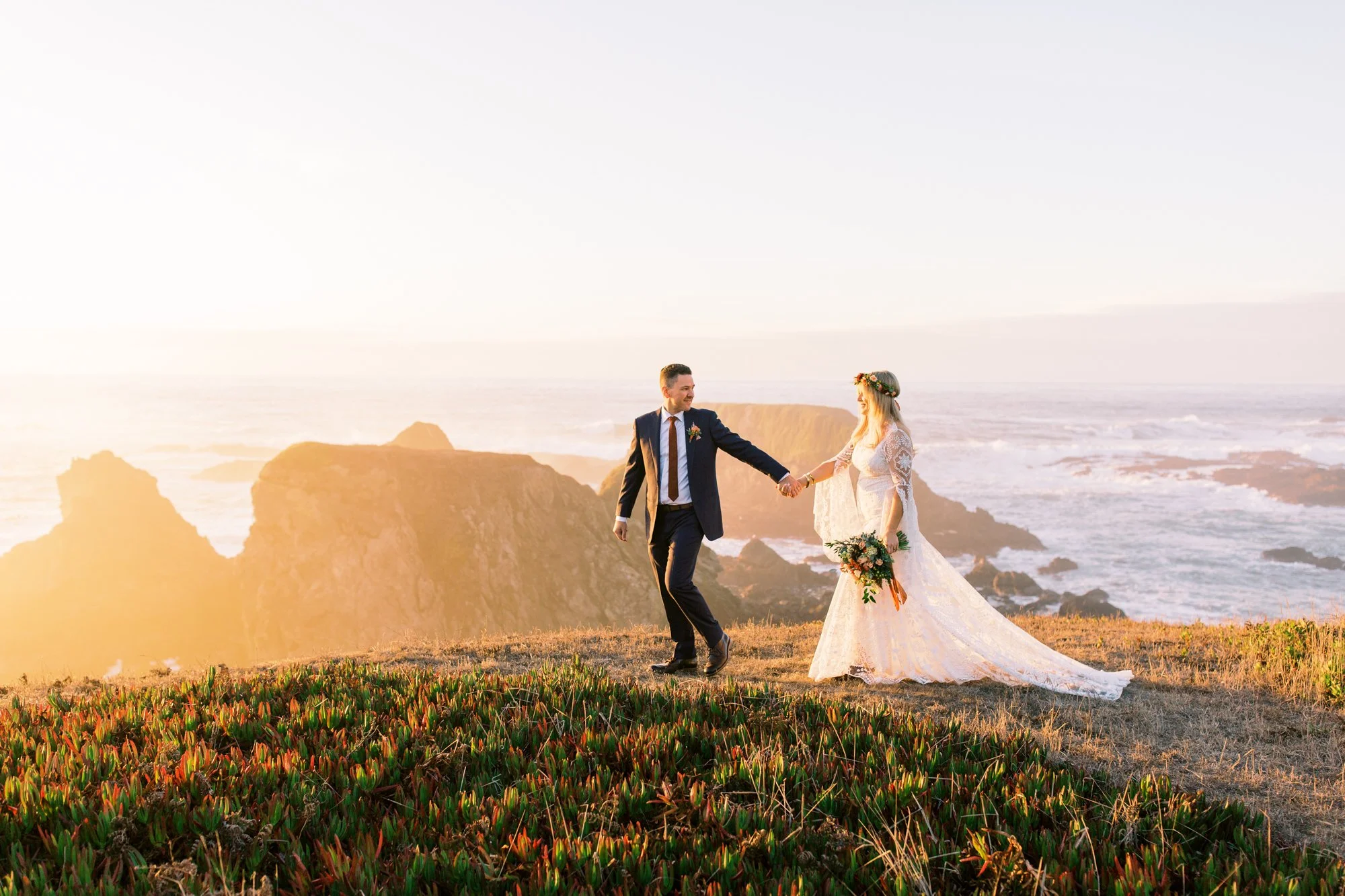 A bride and groom holding hands on a grassy cliffside with ocean waves and rock formations in the background during sunset.