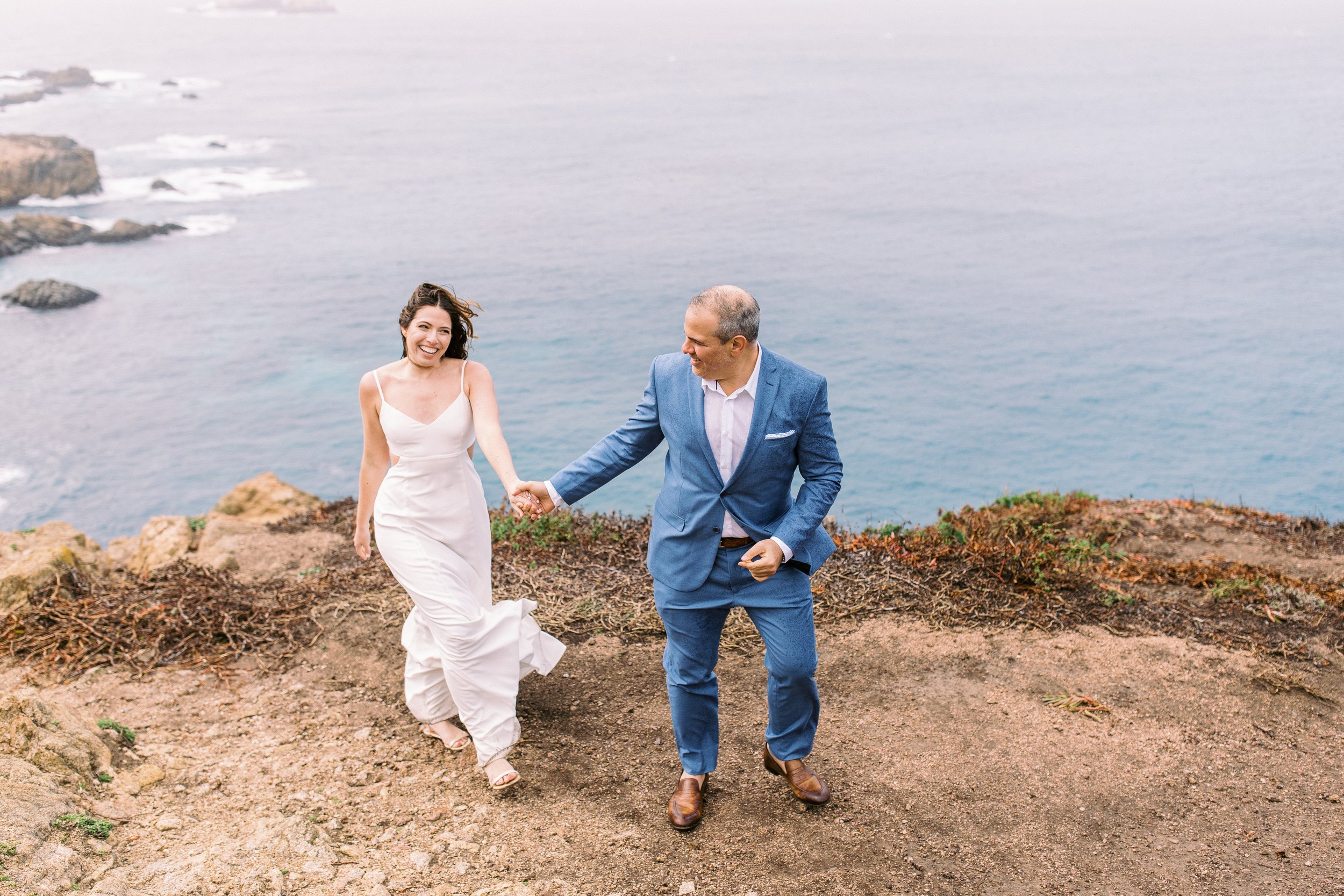 A woman in a white dress and a man in a blue suit hold hands and walk along a cliffside with the ocean in the background.