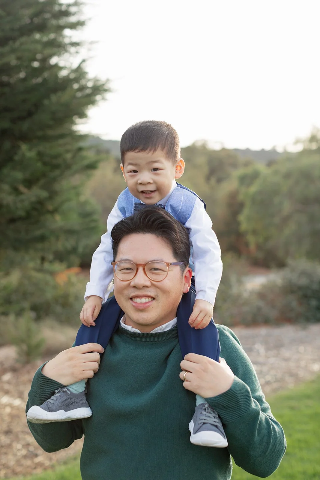 A man carrying a young boy on his shoulders outdoors in a park with trees in the background.