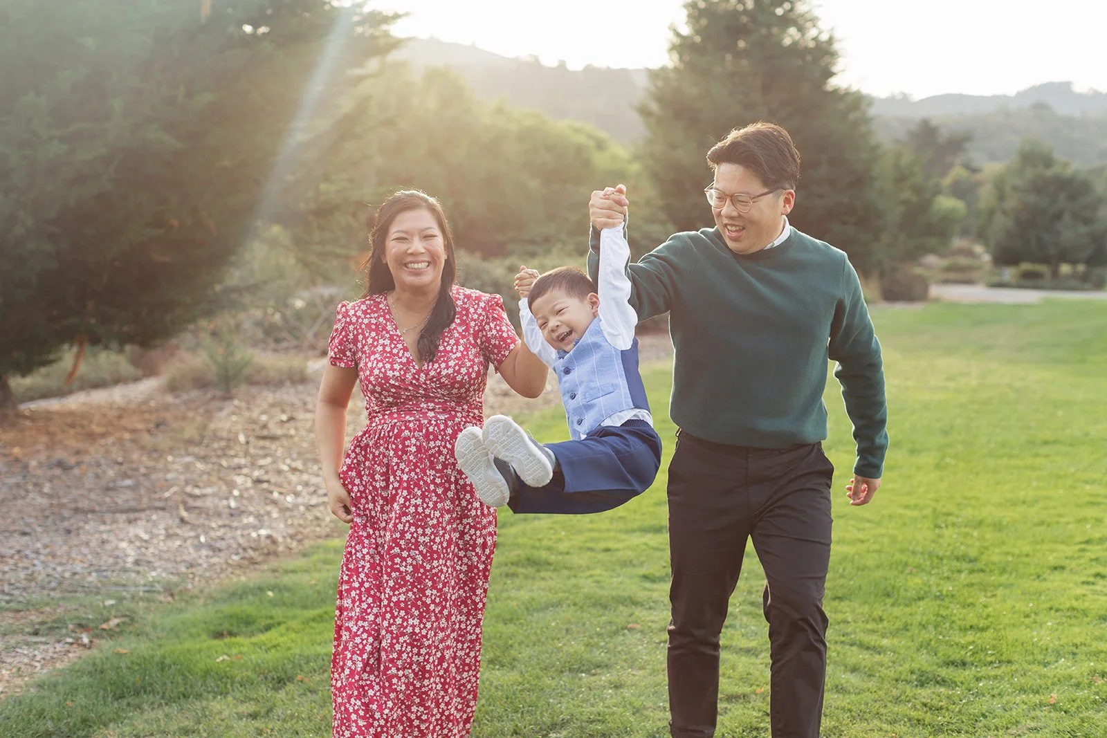A family of three enjoying a moment outdoors, with the man lifting a young boy off the ground while the woman watches and smiles, all set in a park with trees and greenery in the background.