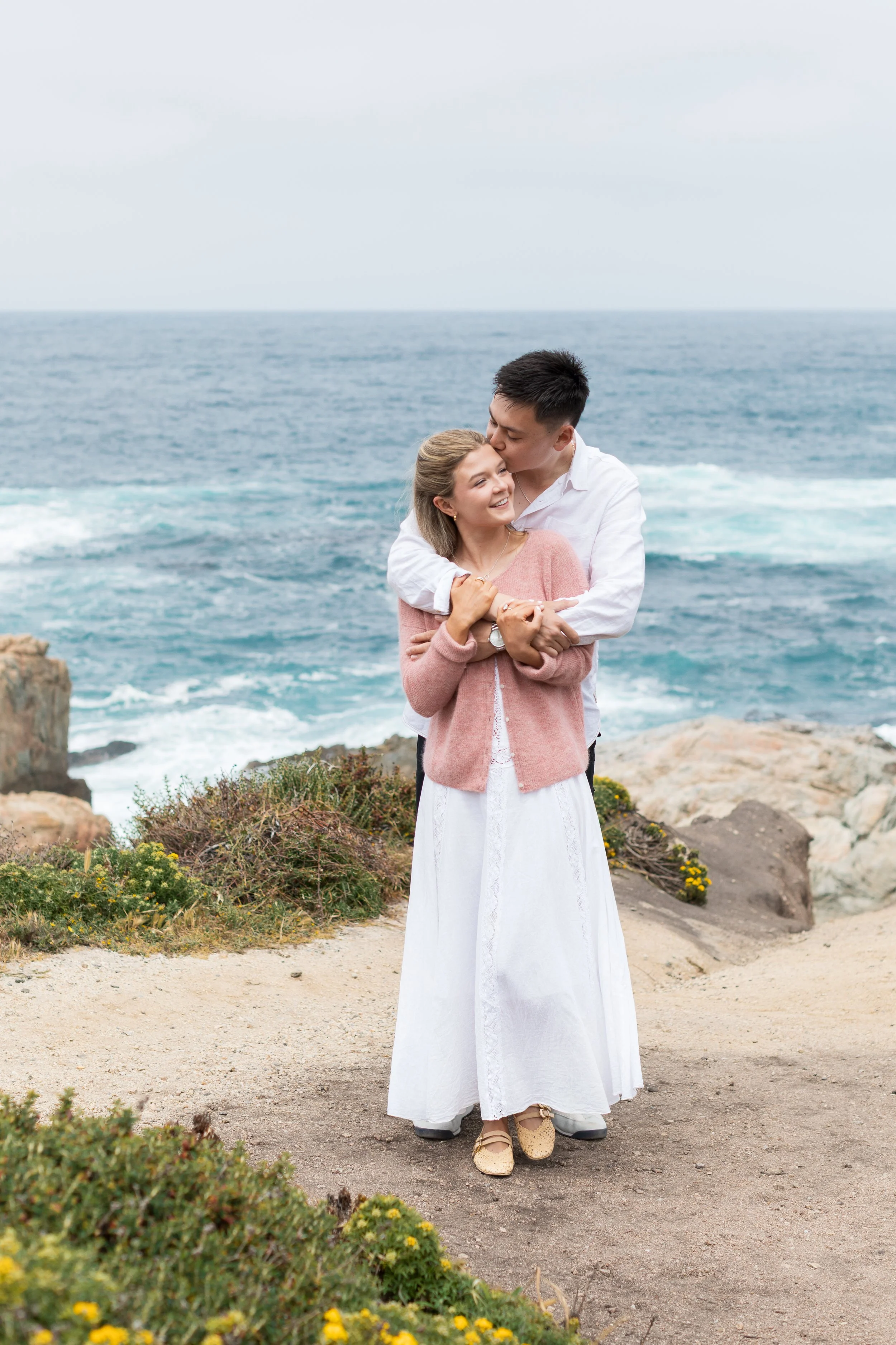 A couple embracing on a rocky coastal area, with the ocean and sky in the background. The man is giving the woman a kiss on the forehead while she is smiling, and they appear happy.