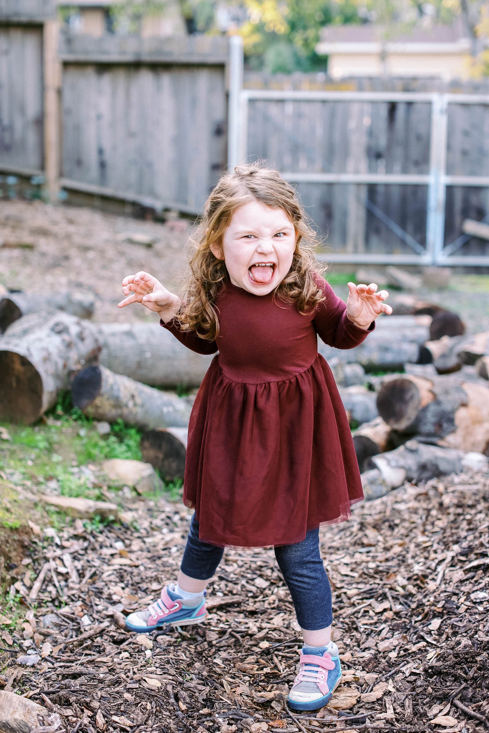 A young girl in a maroon dress and blue sneakers with pink laces standing outdoors on a wooded ground, making a fierce face with her mouth open and hands raised as if pretending to be a monster or a superhero.