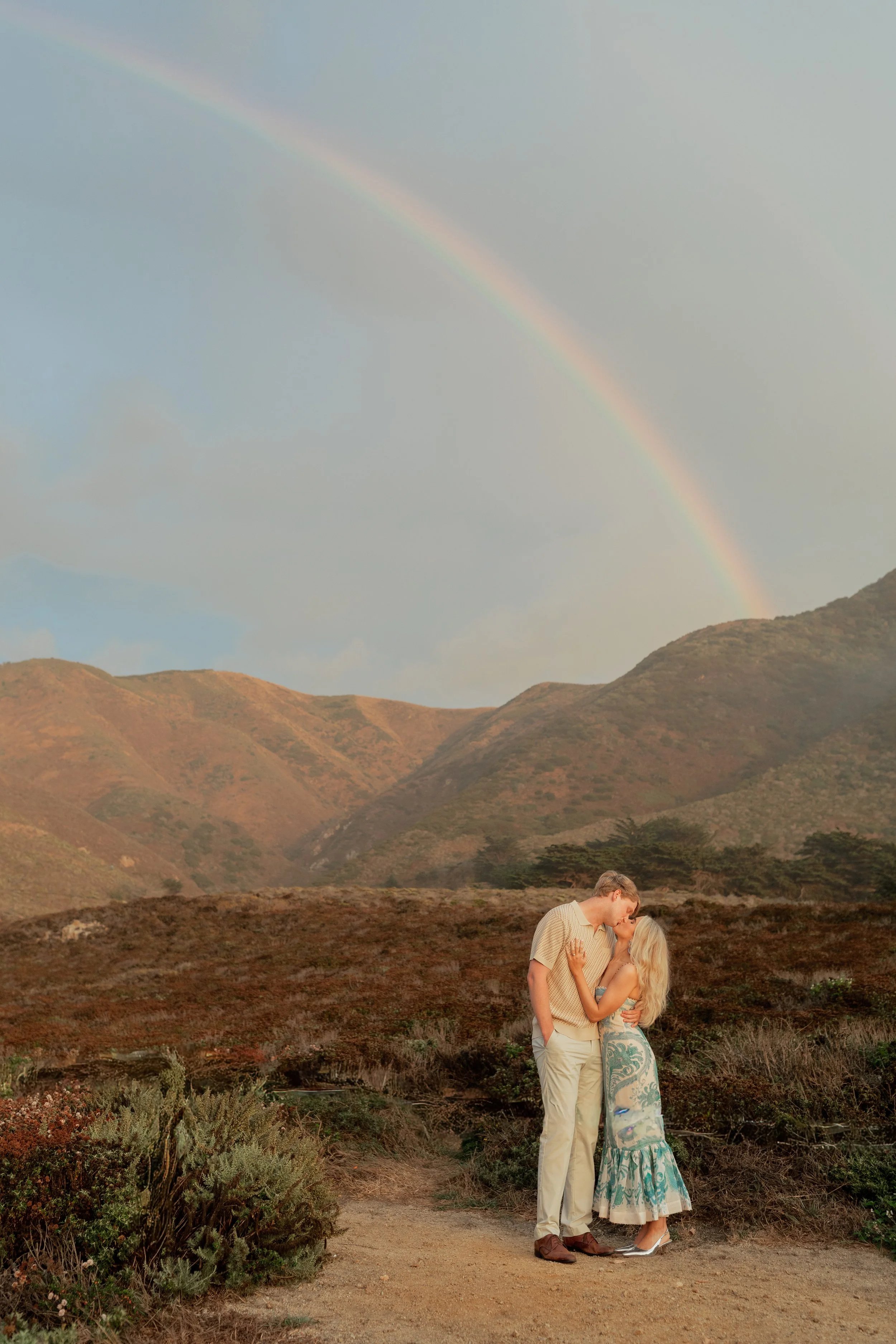 A couple kissing outdoors on a dirt path with mountain hills and a rainbow in the sky.