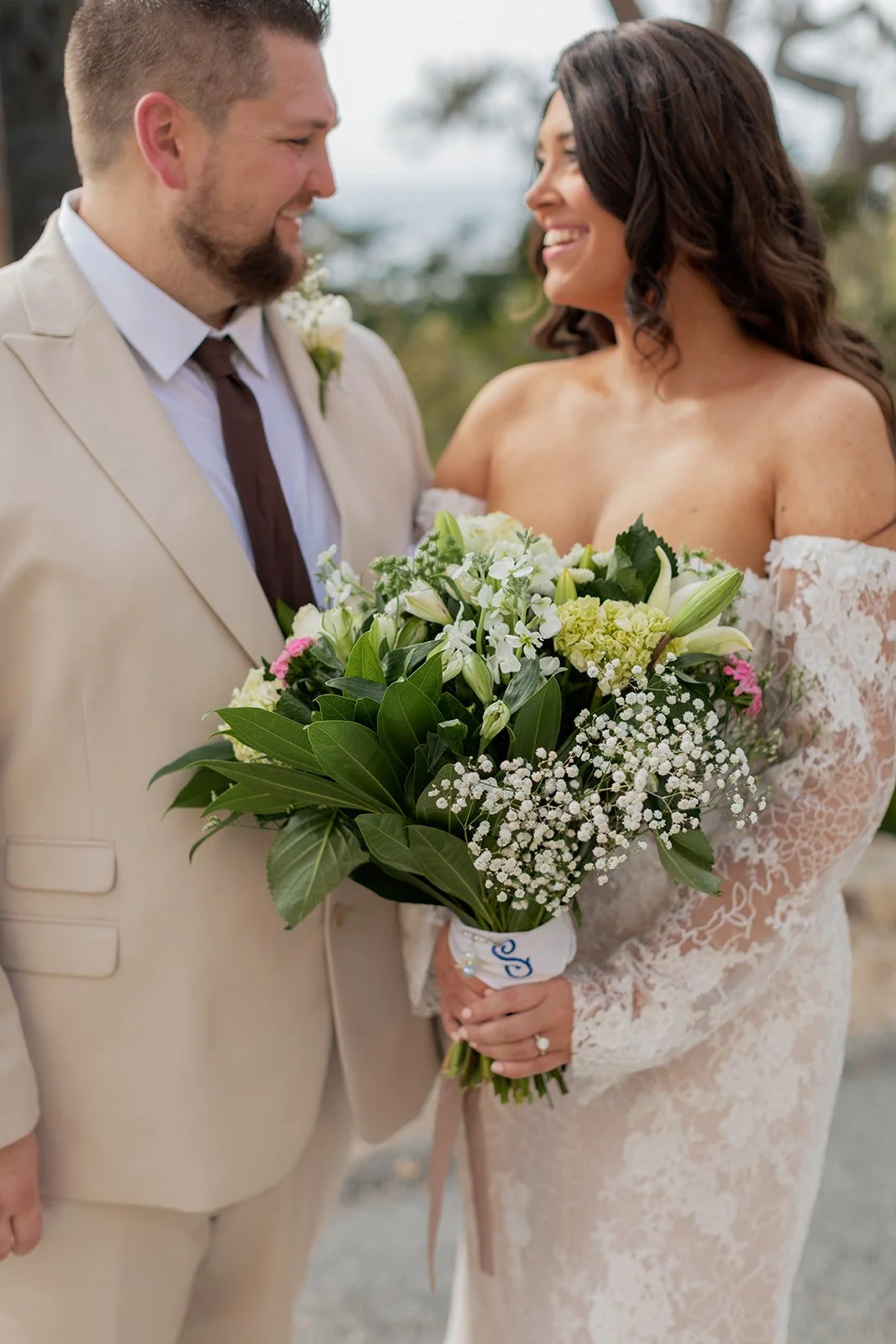 Newlywed couple smiling at each other, holding a bouquet of white flowers, outdoors.