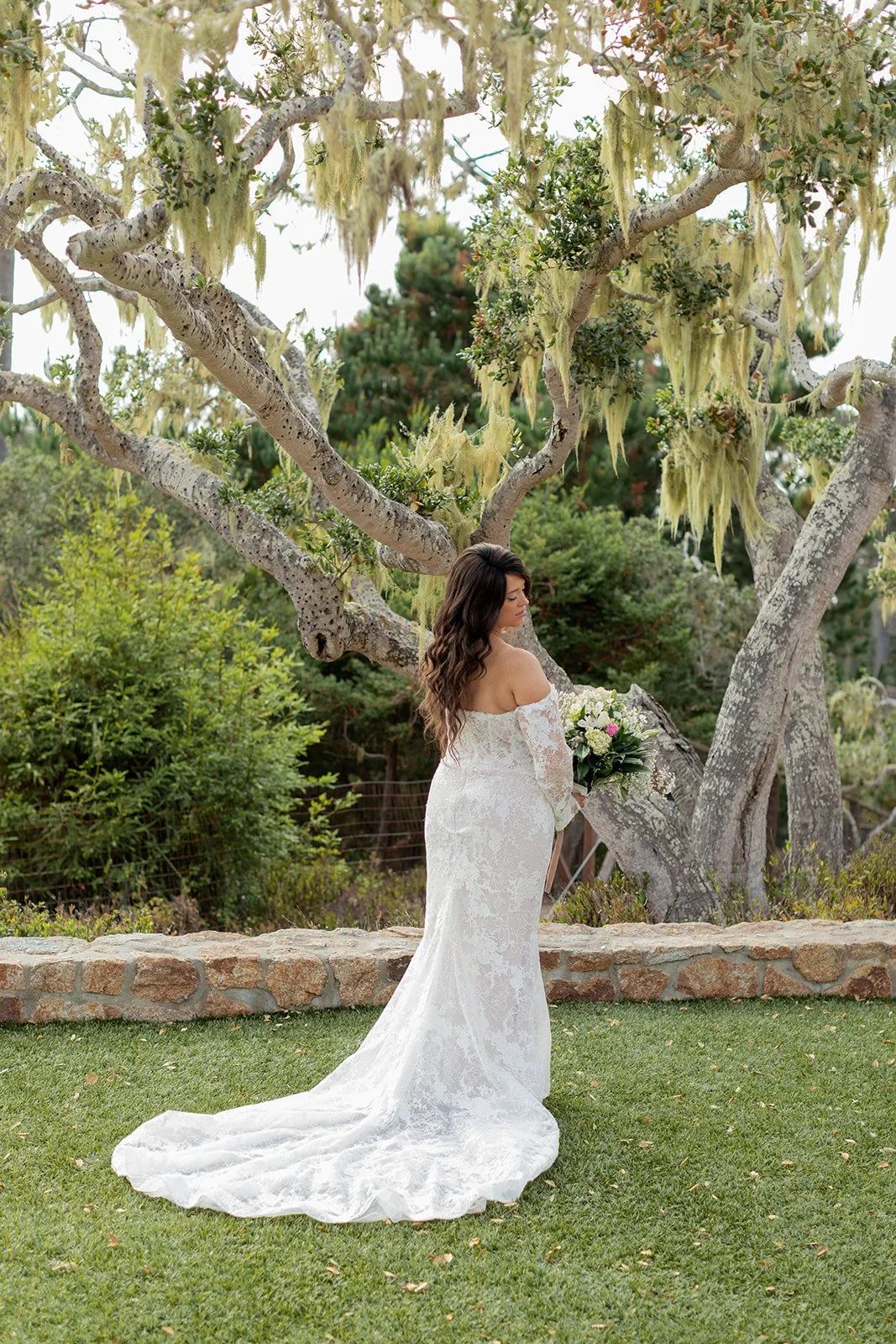 Brunette woman in a white lace off-shoulder wedding dress standing outdoors on grass, holding a bouquet, near a gnarled tree with hanging moss and green foliage.
