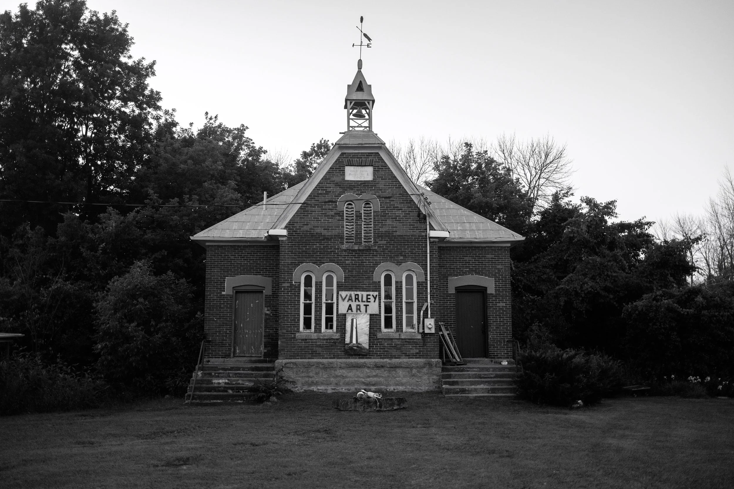 A small brick church with a bell tower and a weather vane on top, surrounded by trees, with steps leading up to the entrance and a sign that says "Varley Art" in front.