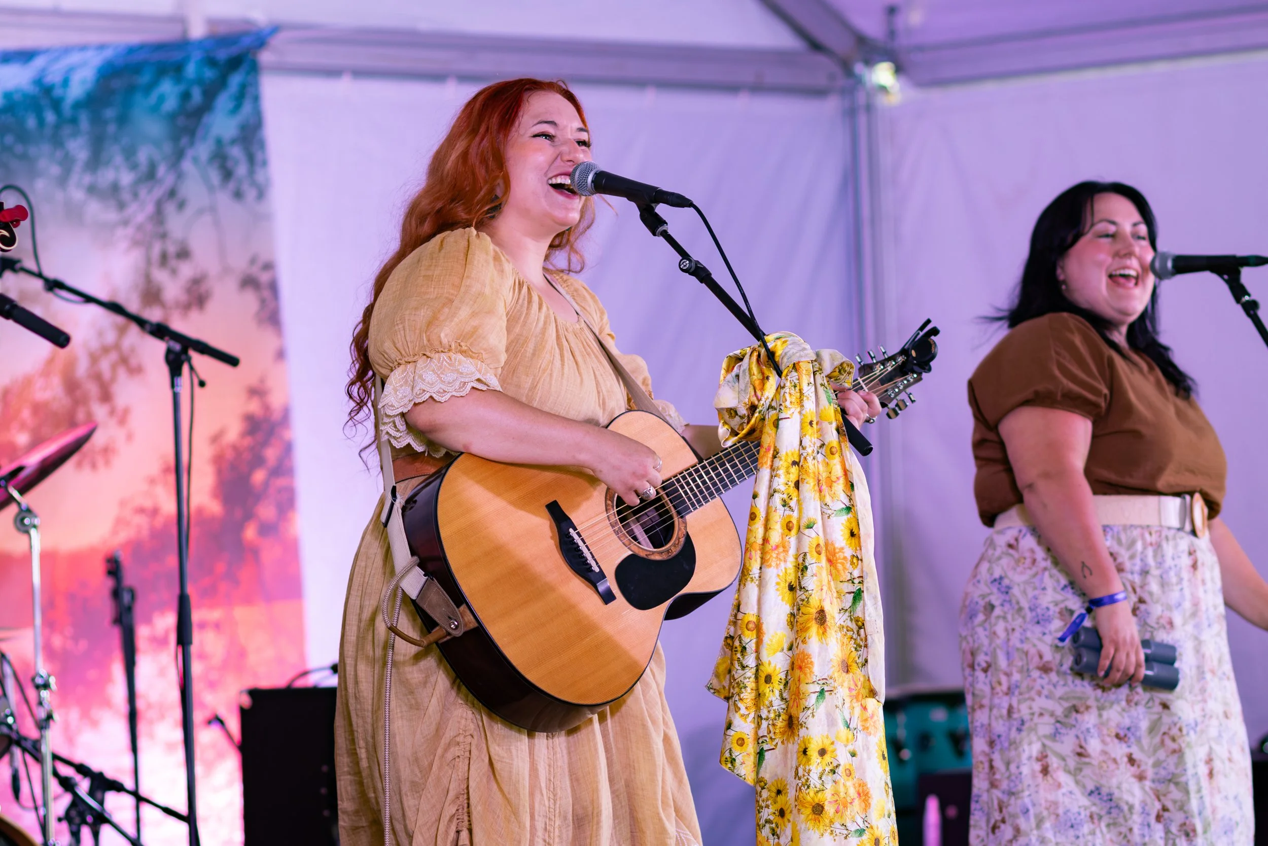 Two women singing and playing guitar on stage at an outdoor event, with microphones and musical equipment around them.