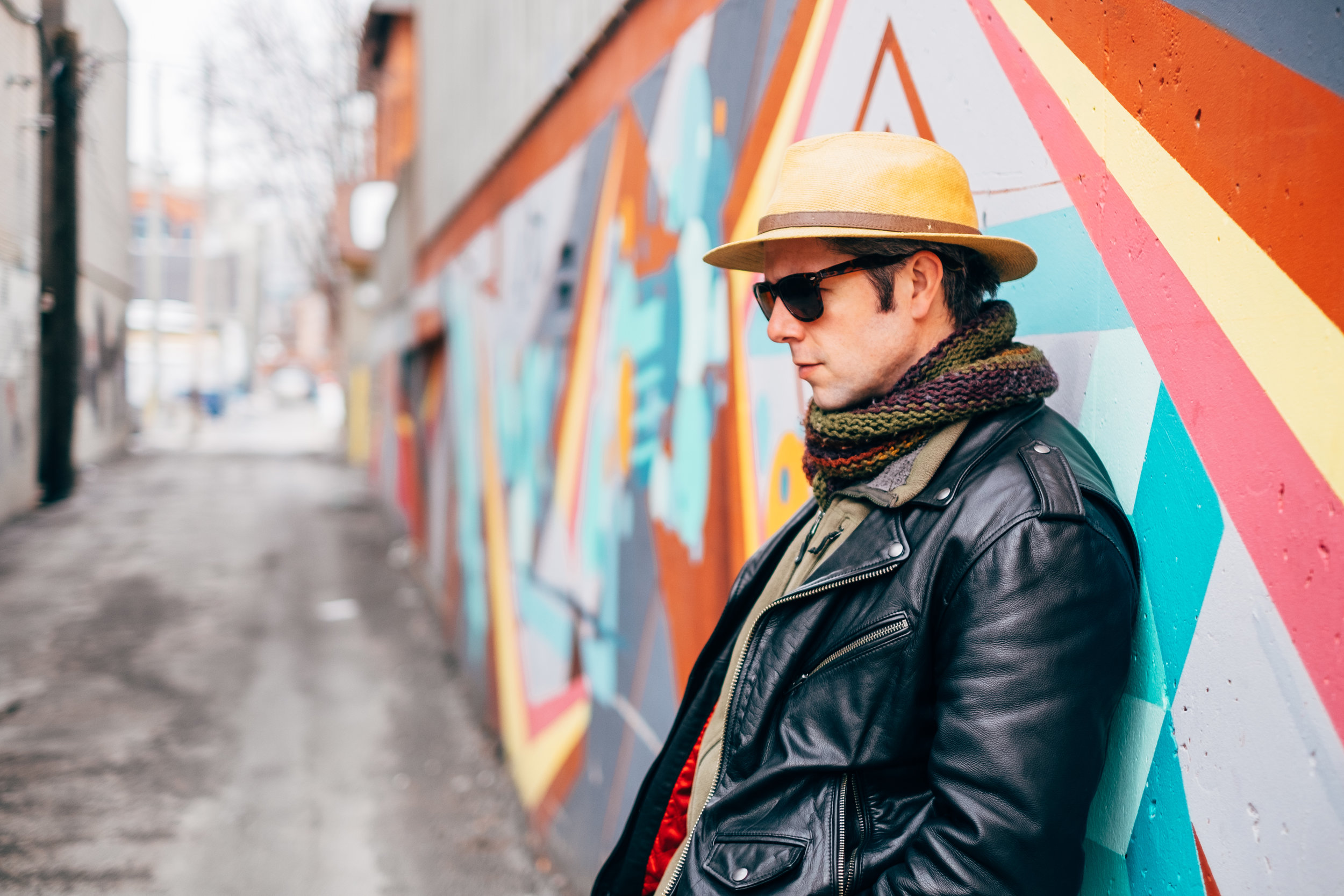 A man in sunglasses, a hat, a scarf, and a leather jacket leaning against a colorful graffiti wall in an urban alley.