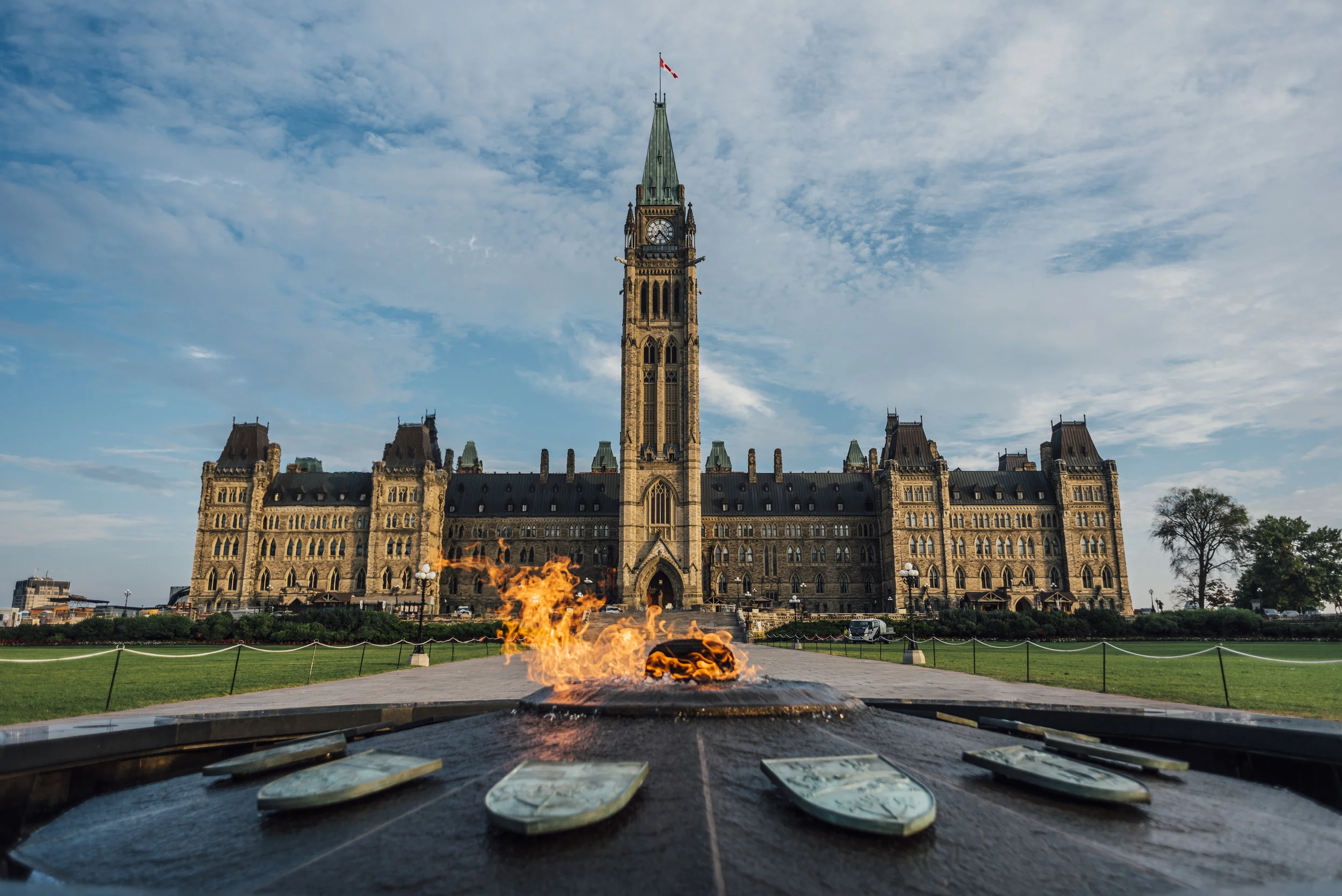 Parliament Hill with the Peace Tower in Ottawa, Canada, in daytime with a clear sky and an eternal flame memorial in the foreground.