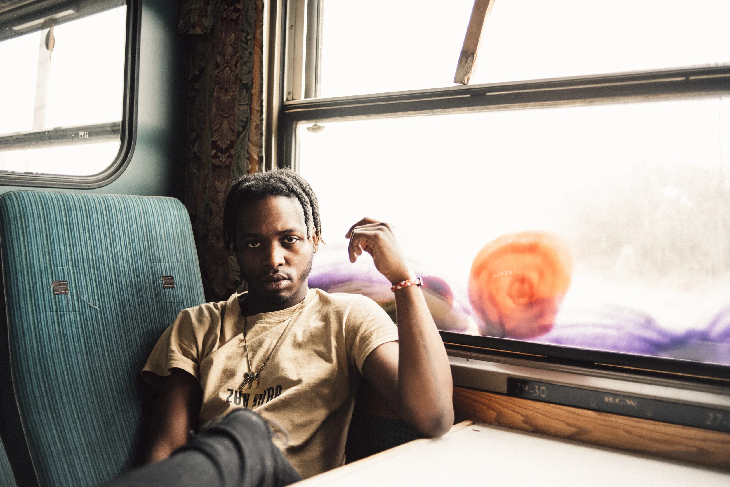 A young man sitting on a train with a serious expression, wearing a beige T-shirt, near a window with a colorful abstract design.