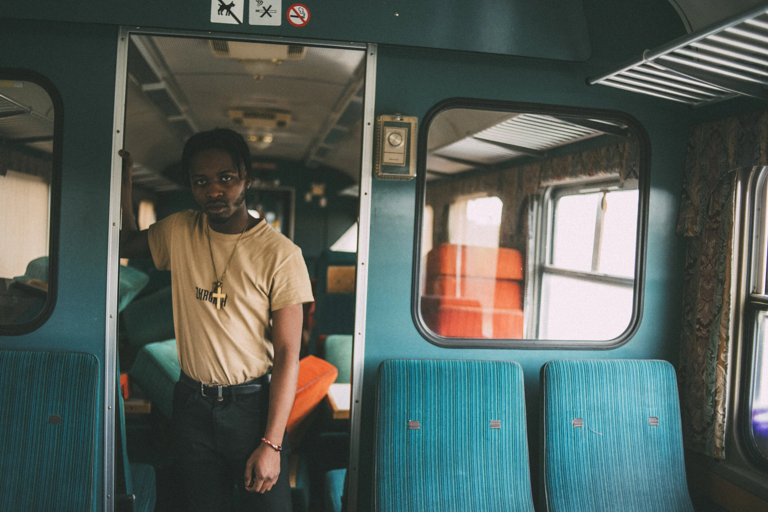 A young man with dreadlocks, wearing a beige T-shirt with a necklace, standing inside a vintage bus with blue seats, windows, and curtains, looking at the camera.