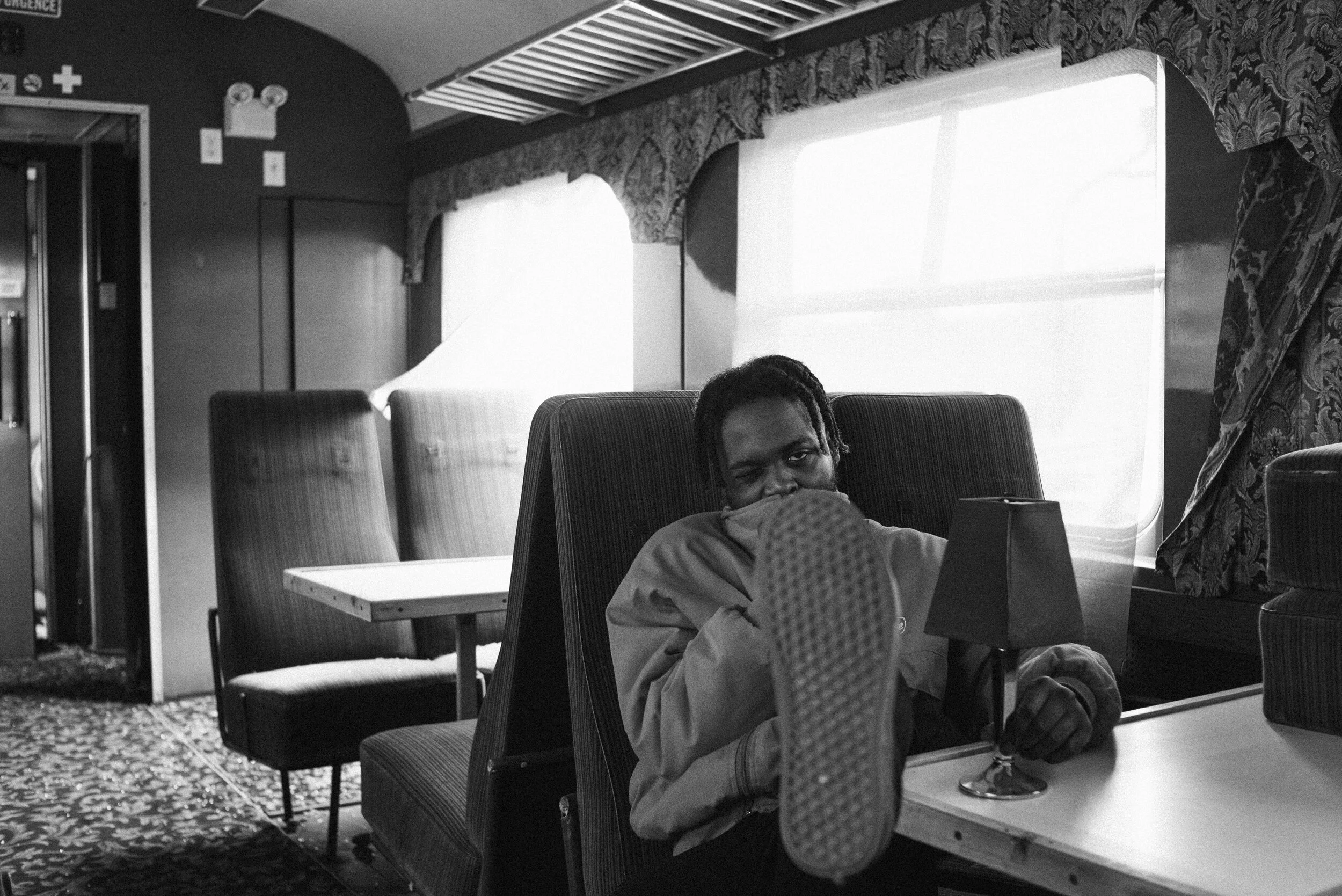 A man sitting in a cafe or restaurant, with his foot resting on a table and looking at the camera. The setting has upholstered chairs, windows with curtains, and a patterned carpet.