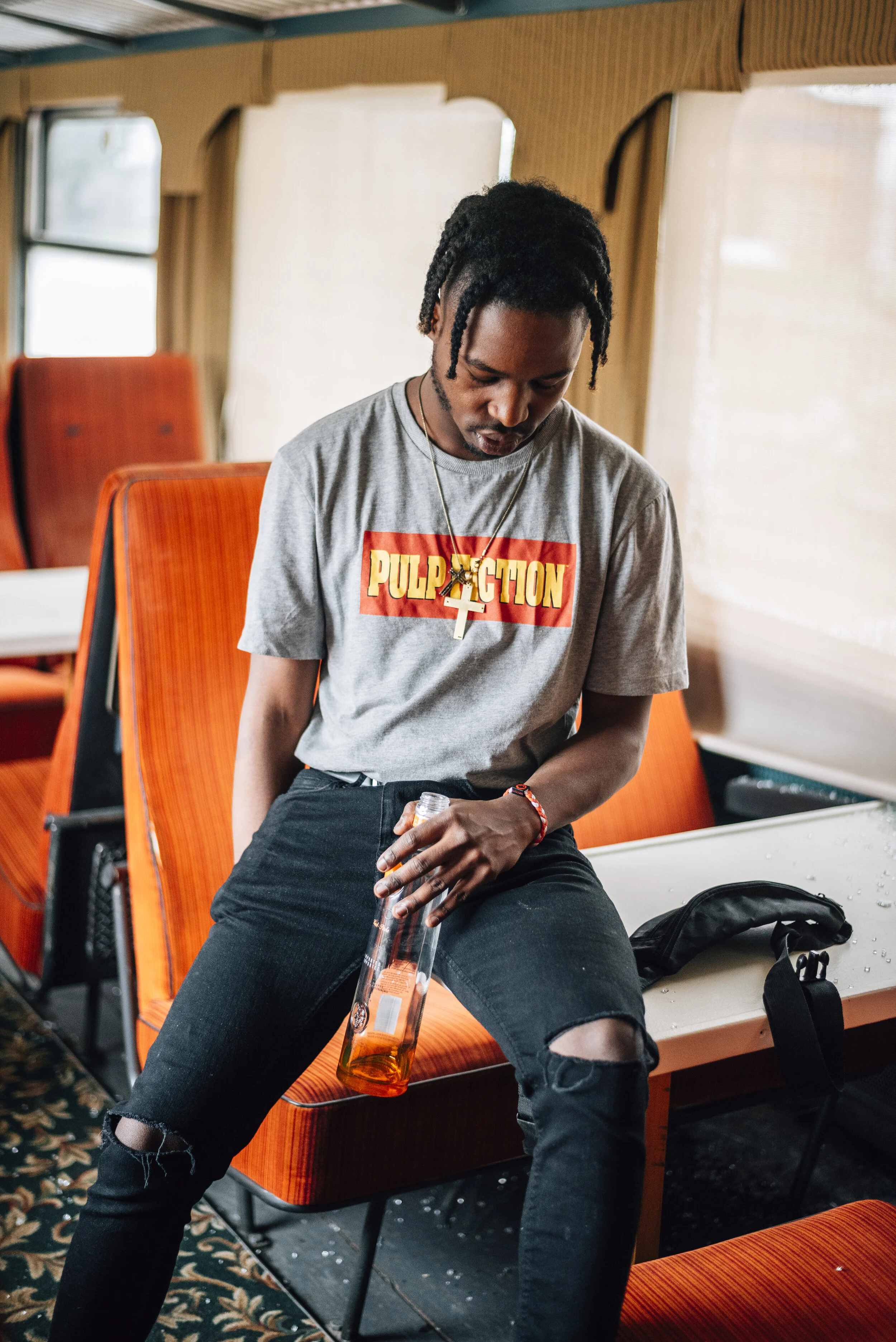 A young man with dreadlocks wearing a 'Pulp Fiction' t-shirt, black ripped jeans, and a necklace, sitting on an orange booth seat in a retro-style room, holding a clear bottle.