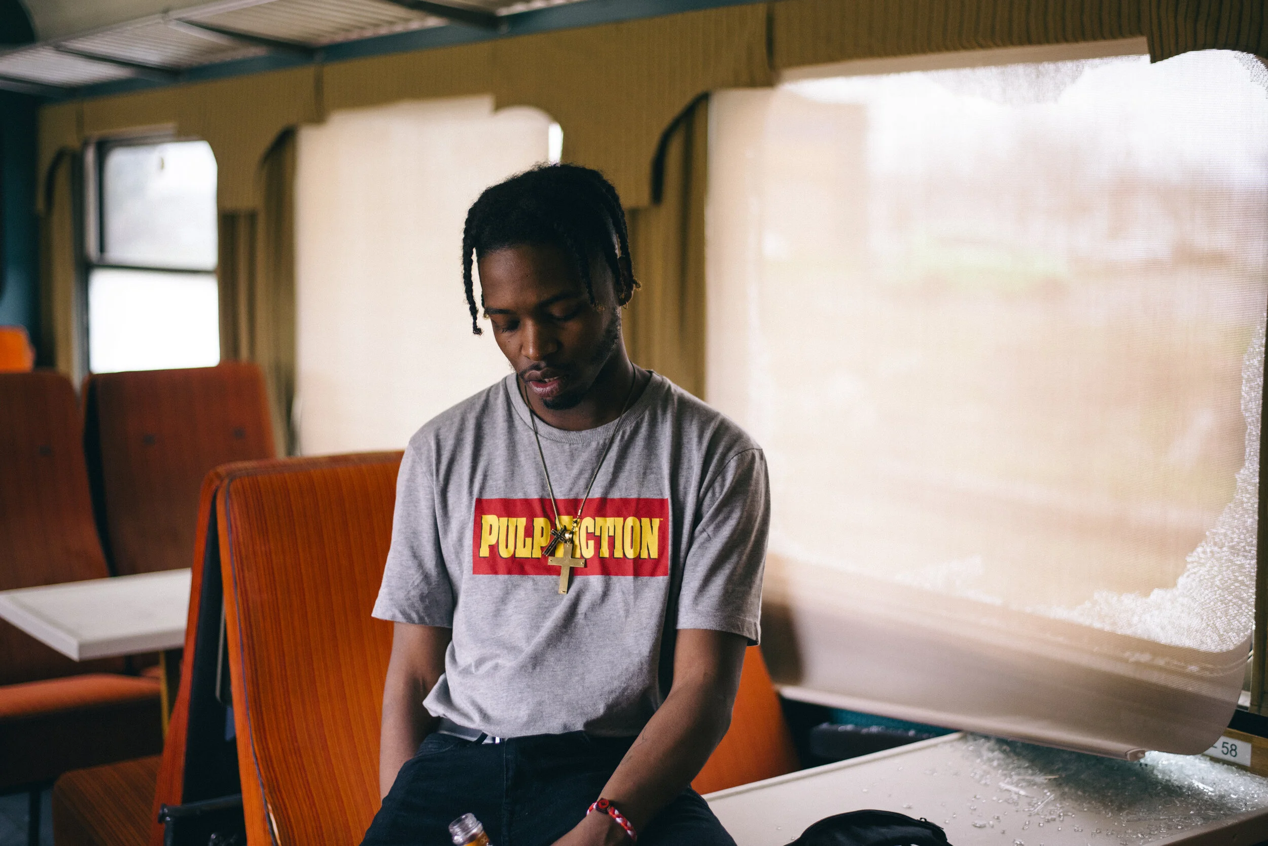A young man with dreadlocks wearing a gray T-shirt with the text 'Pulp Fiction' stands inside a bus with orange seats and broken window glass.