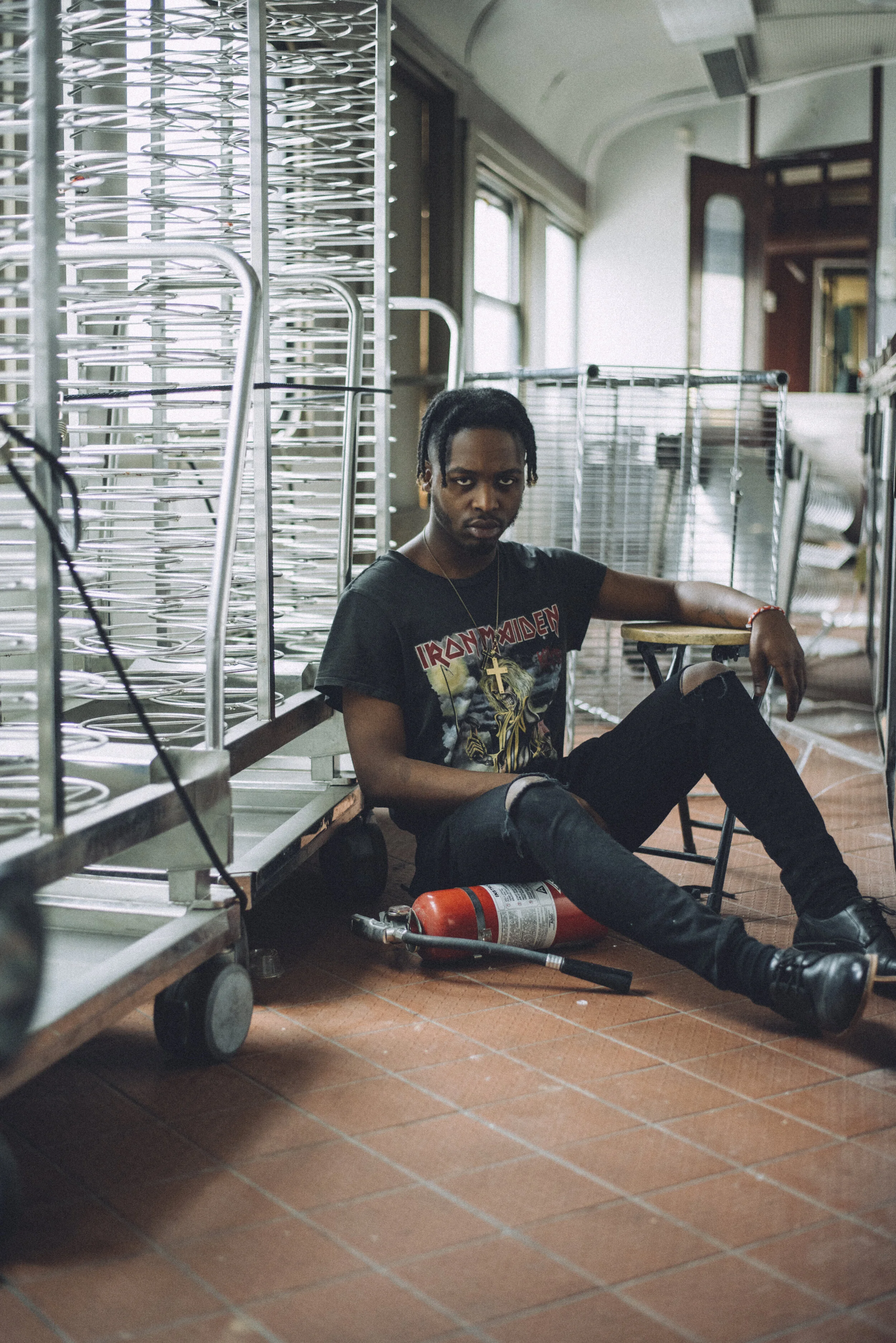 A man with medium-dark skin, dreadlocks, wearing a black Iron Maiden t-shirt, ripped black jeans, and black boots, sitting on the floor next to a fire extinguisher in a room with metal racks and large windows.