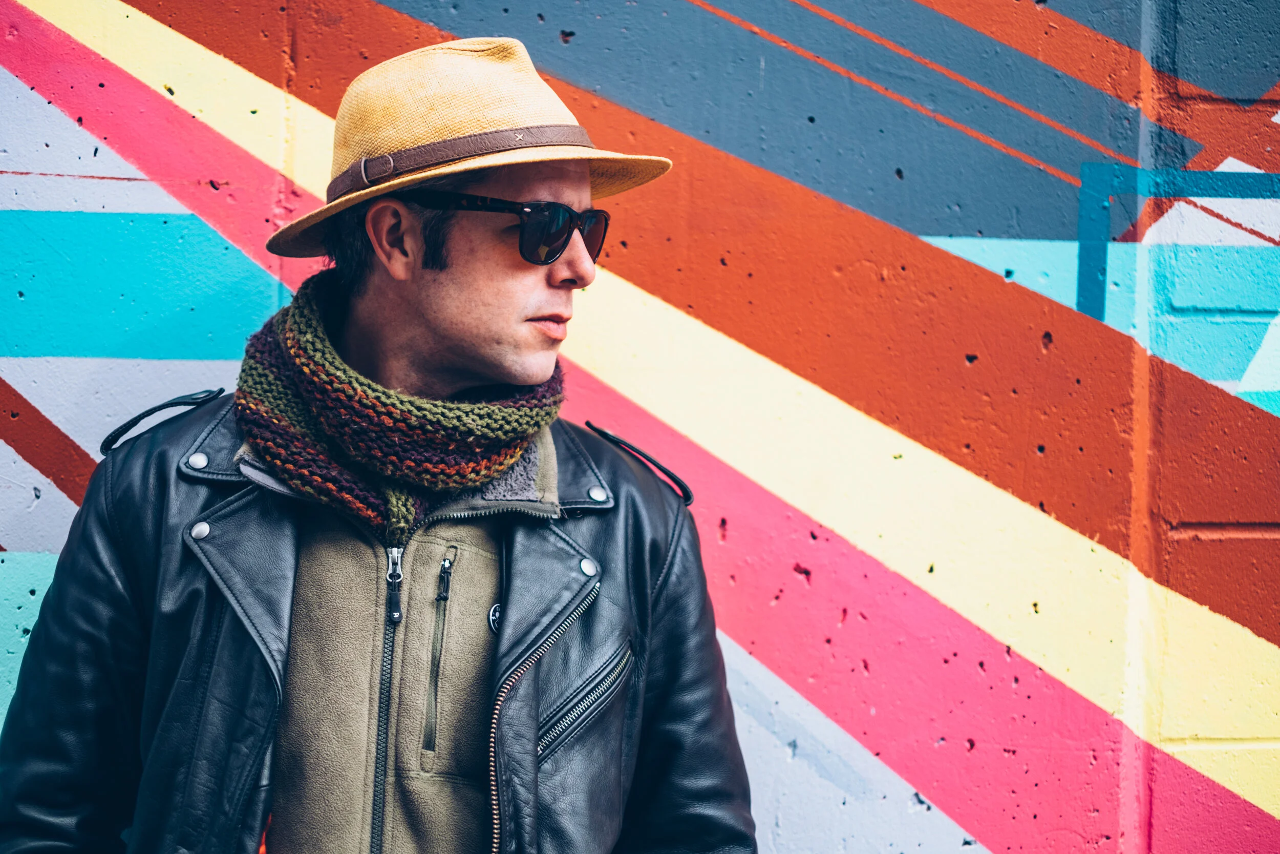A man wearing a tan fedora, black sunglasses, a multicolored scarf, a black leather jacket, and a beige fleece jacket stands in front of a colorful geometric mural painted on a brick wall.