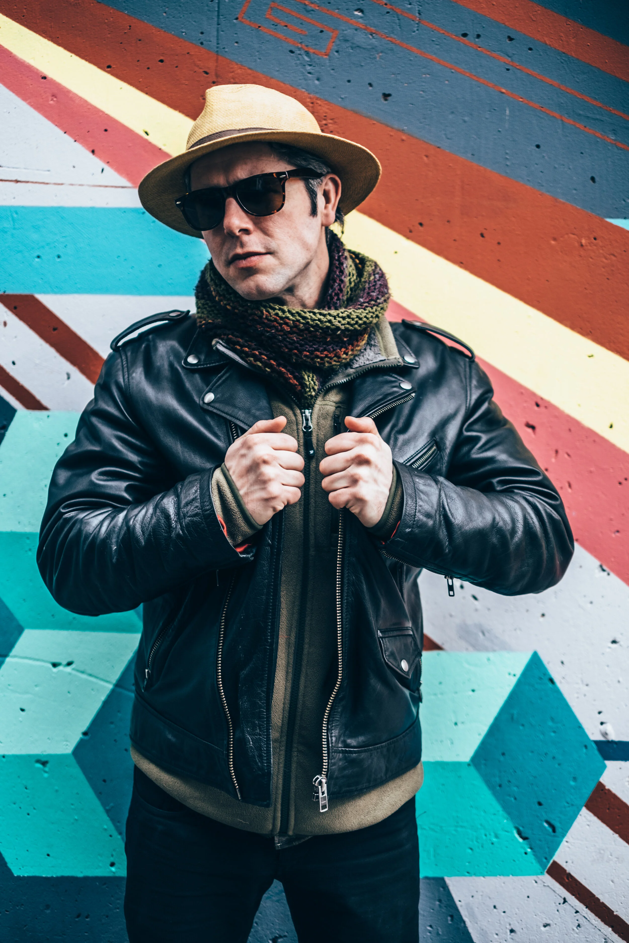 A man wearing a beige fedora, black sunglasses, a colorful scarf, a black leather jacket, and a green hoodie stands in front of a colorful geometric mural wall.