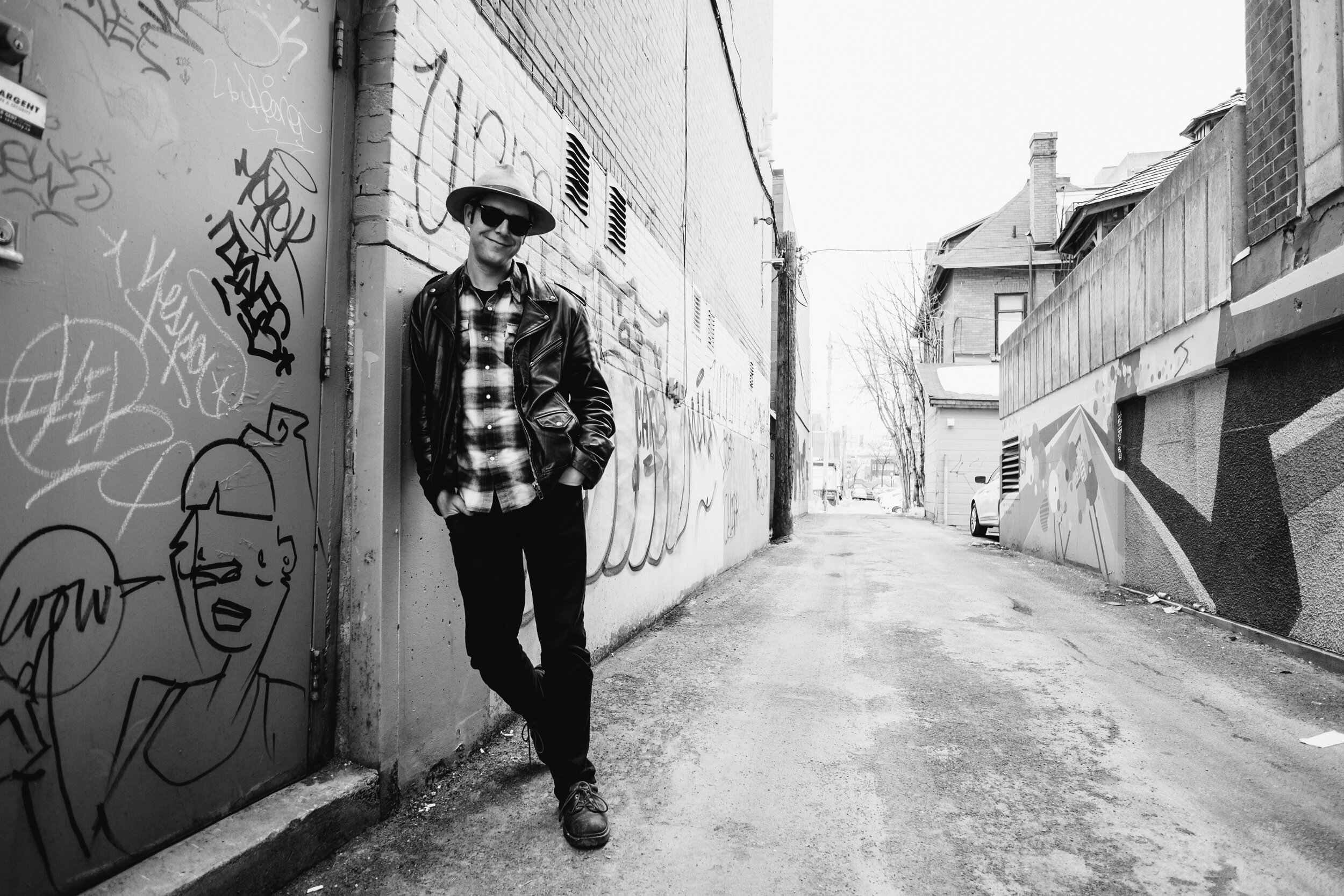 A man wearing sunglasses, a hat, a leather jacket, and a plaid shirt, leaning against a graffiti-covered wall in an alleyway.